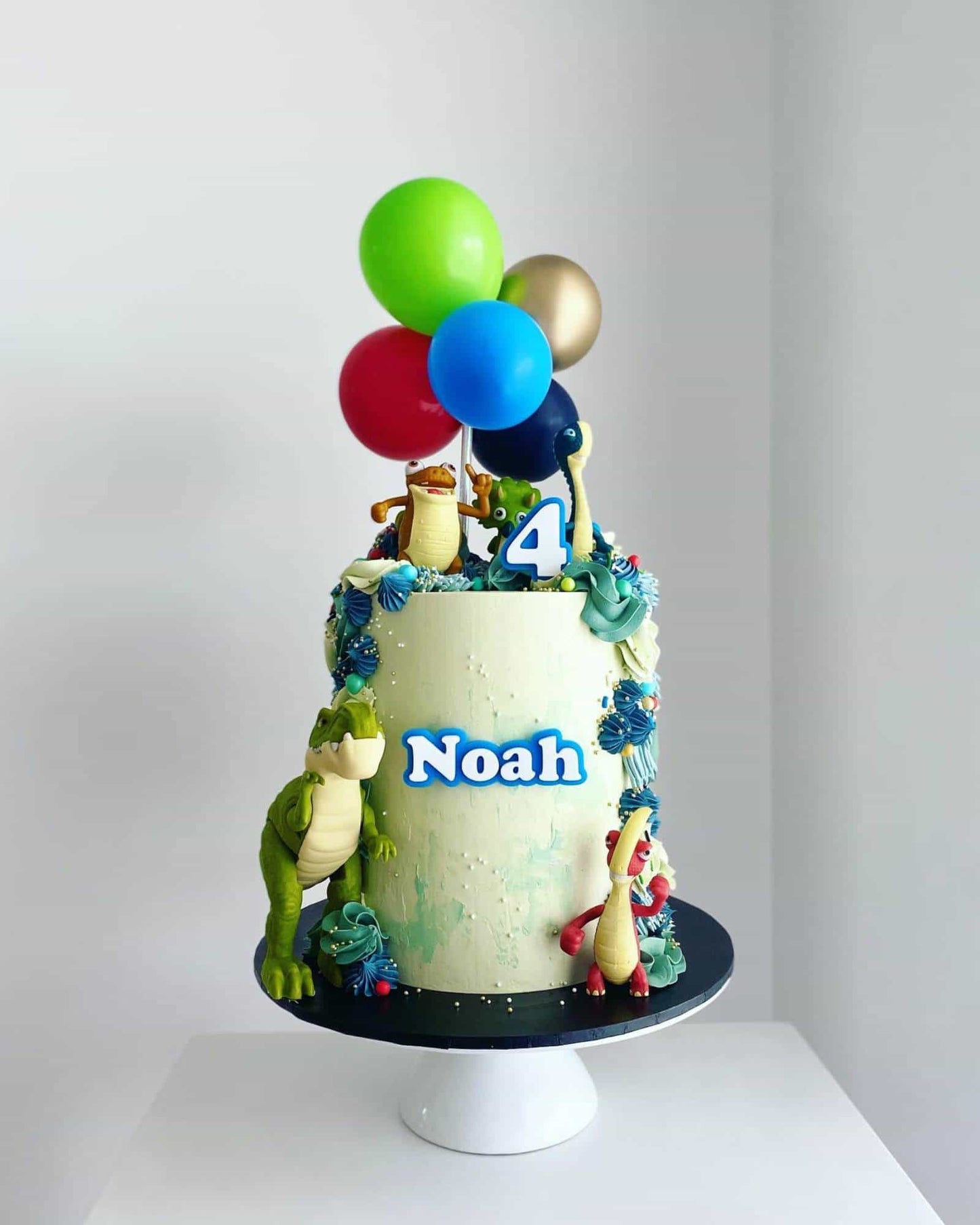 A colorful cake topped with balloons, playful dinosaur figures, and a personalized name for a birthday celebration.