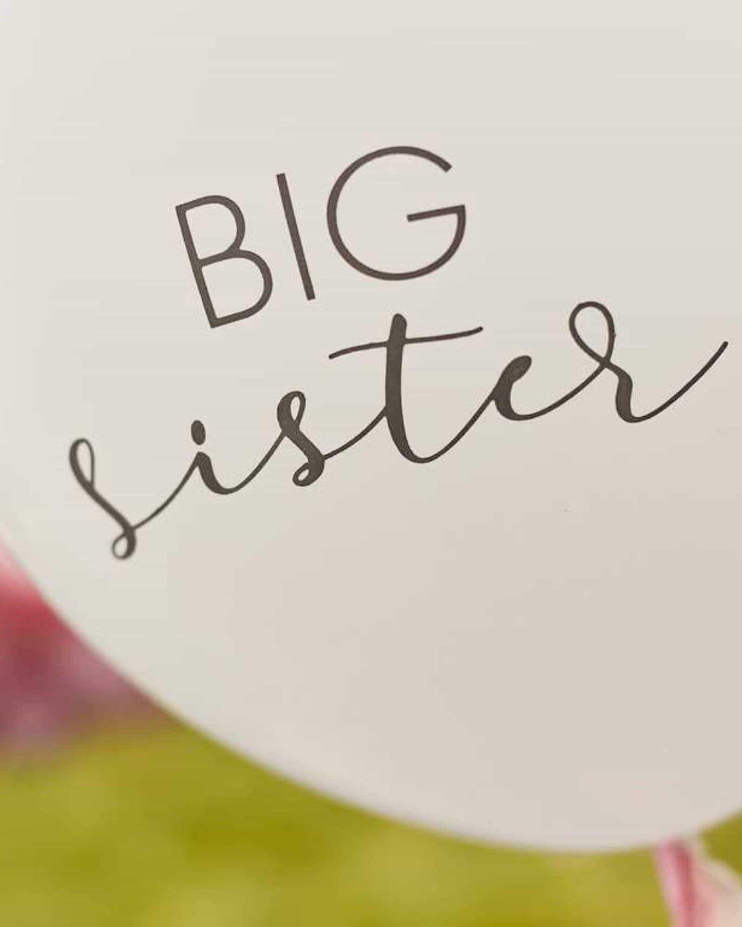 White balloon with "BIG sister" in elegant black script and pink tassels, set against a soft green background.