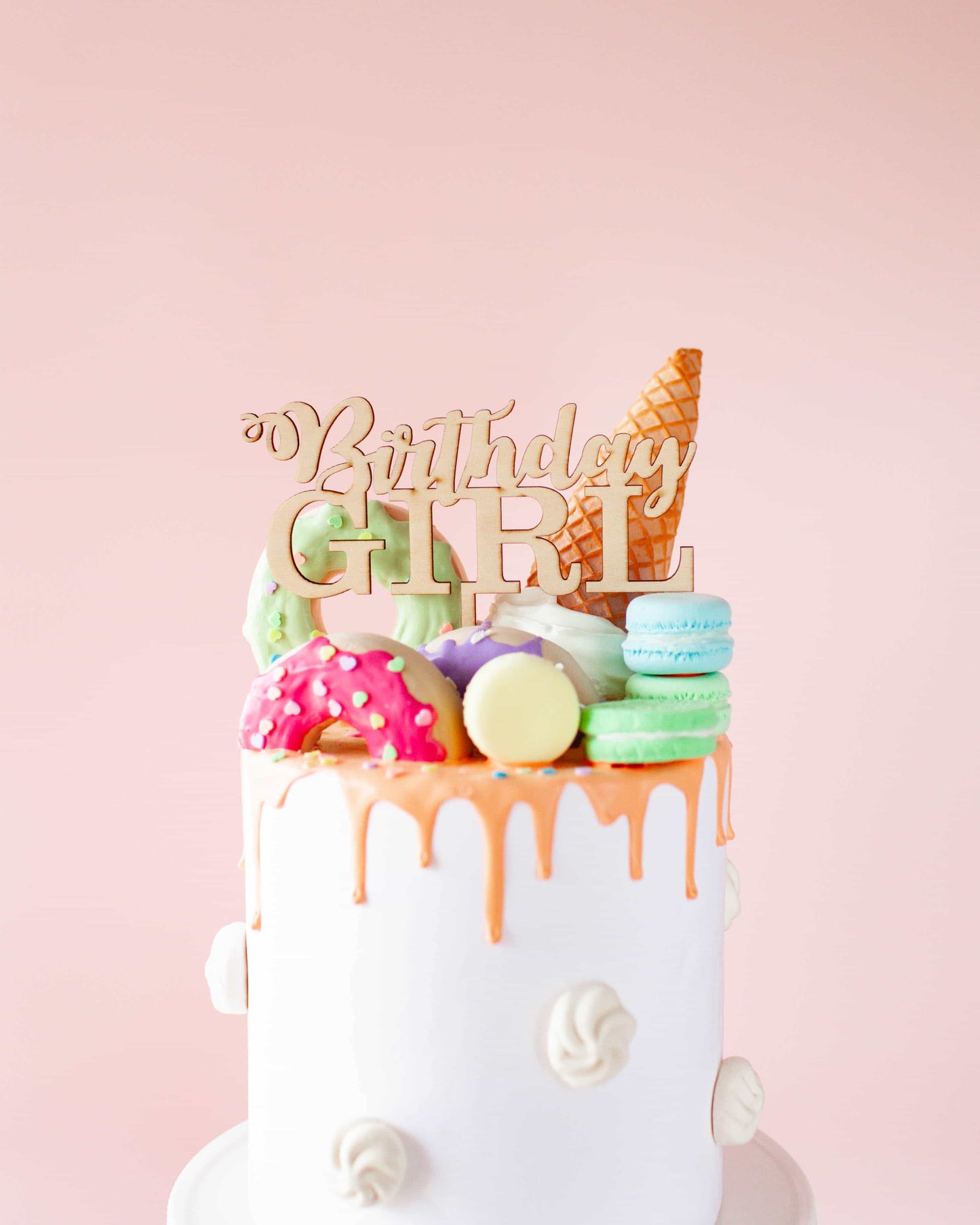 A white cake topped with colorful macarons, donuts, and a wooden "Birthday Girl" topper against a soft pink background.