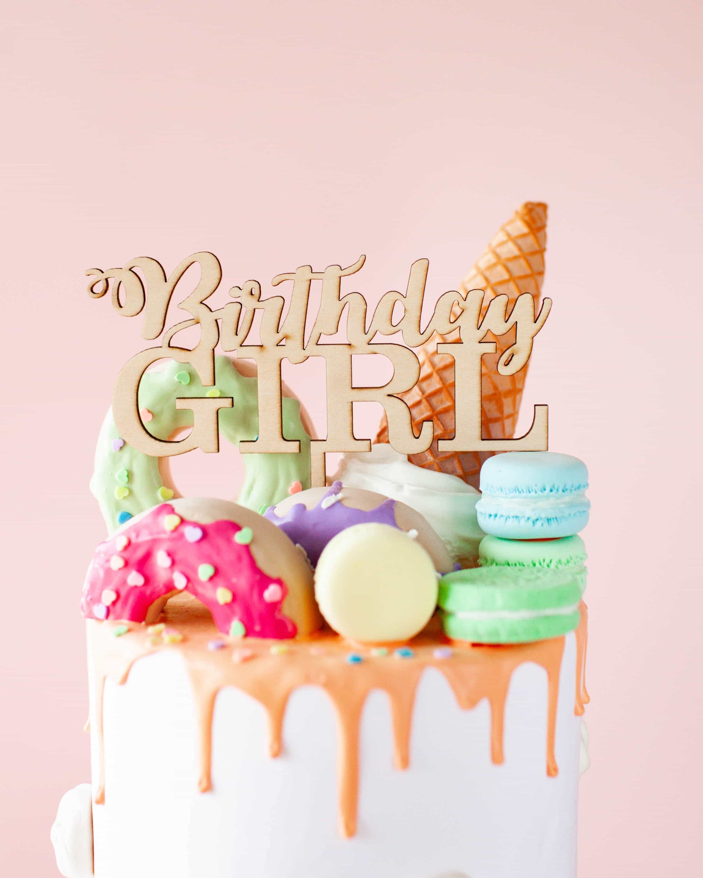 Wooden topper reads "Birthday Girl," surrounded by colorful confections on a cake with a pink background.