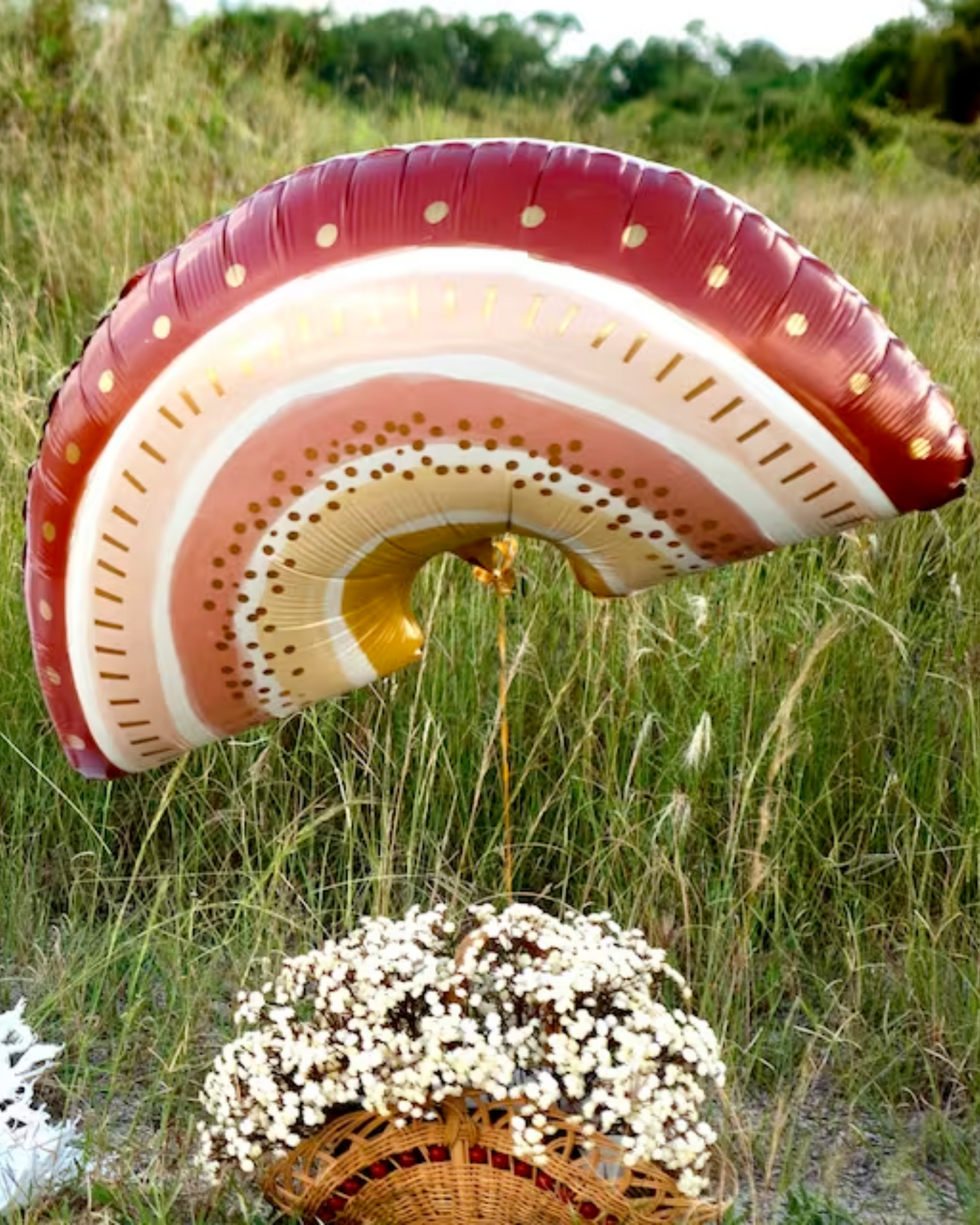 A colorful rainbow-shaped decoration floats above a woven basket filled with delicate white flowers in a grassy field.