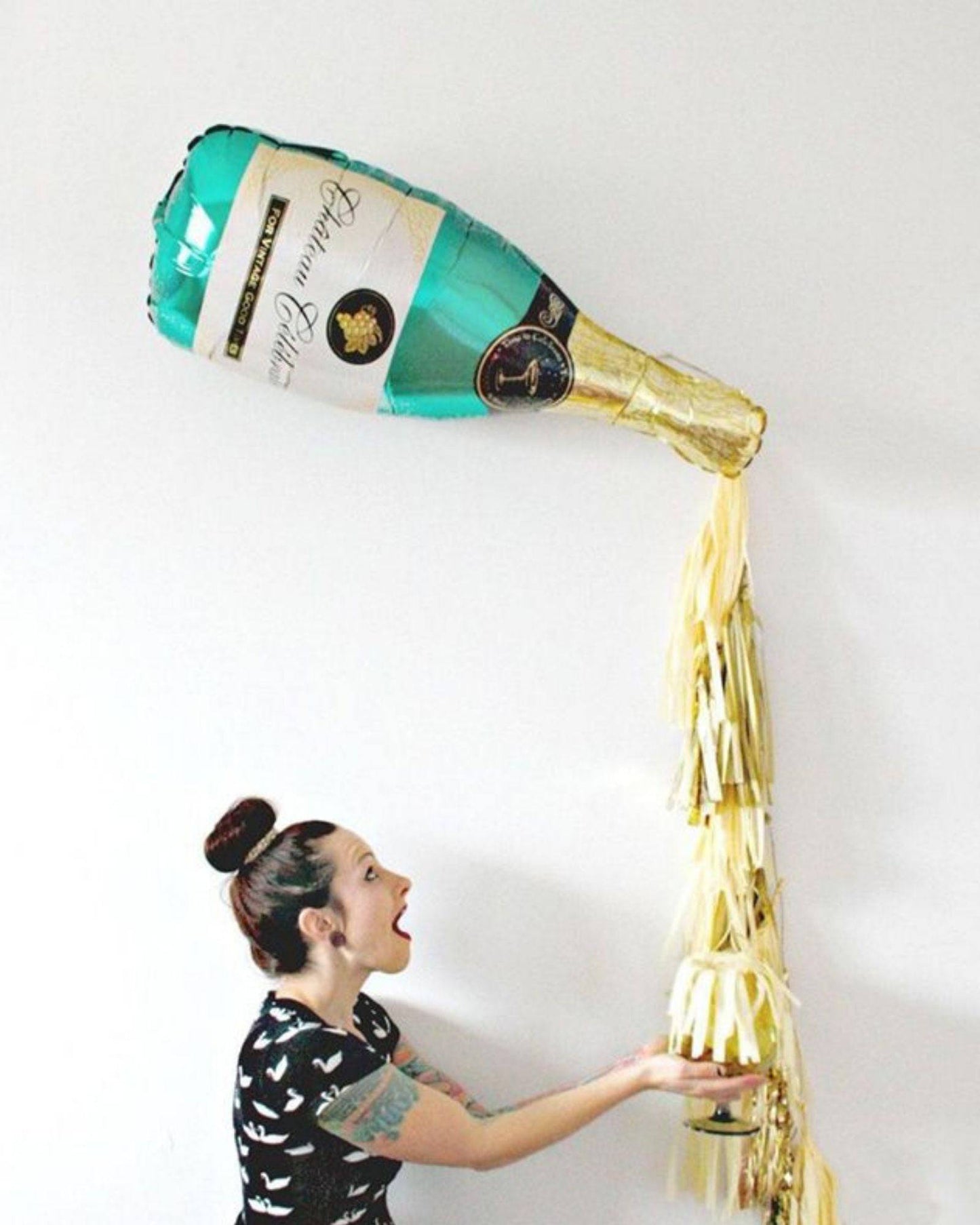 A woman joyfully holds a glass under a large champagne bottle balloon, with golden streamers cascading down.