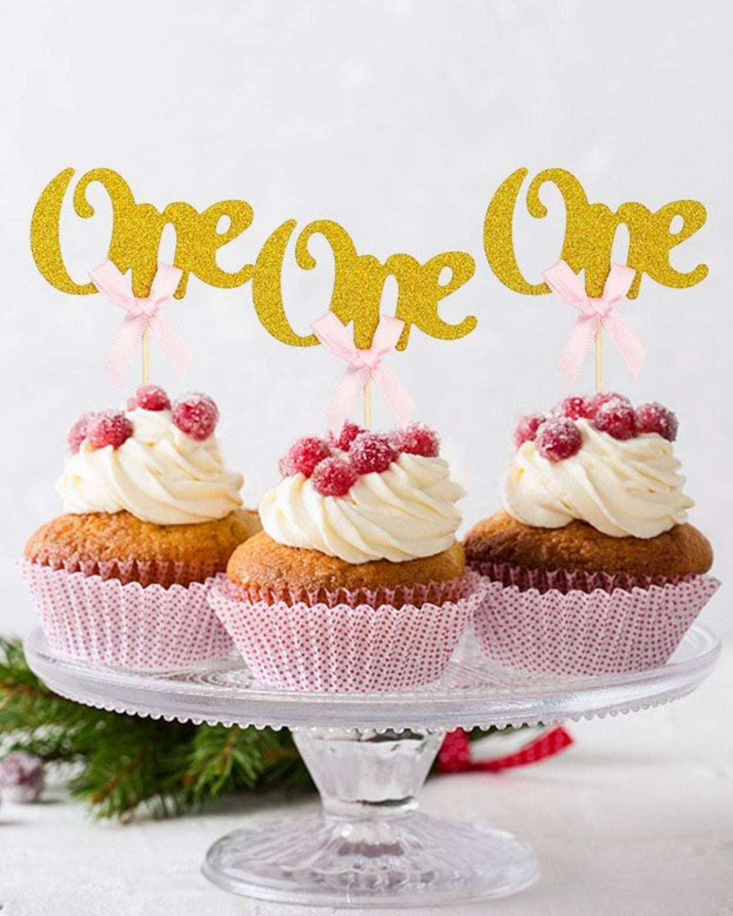 Three cupcakes topped with swirls of frosting, adorned with raspberries and gold "One" picks with pink bows.