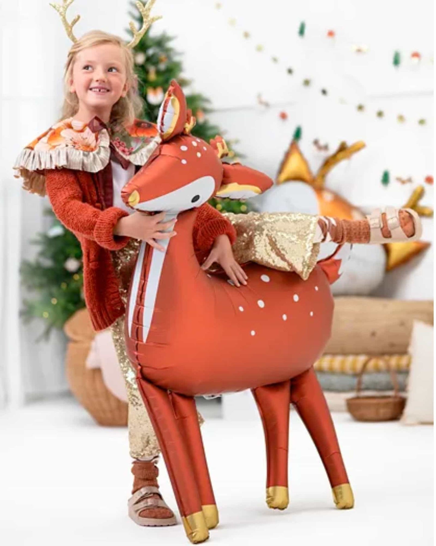 A child in festive attire joyfully interacts with a large, deer-shaped foil balloon in a decorated holiday setting.