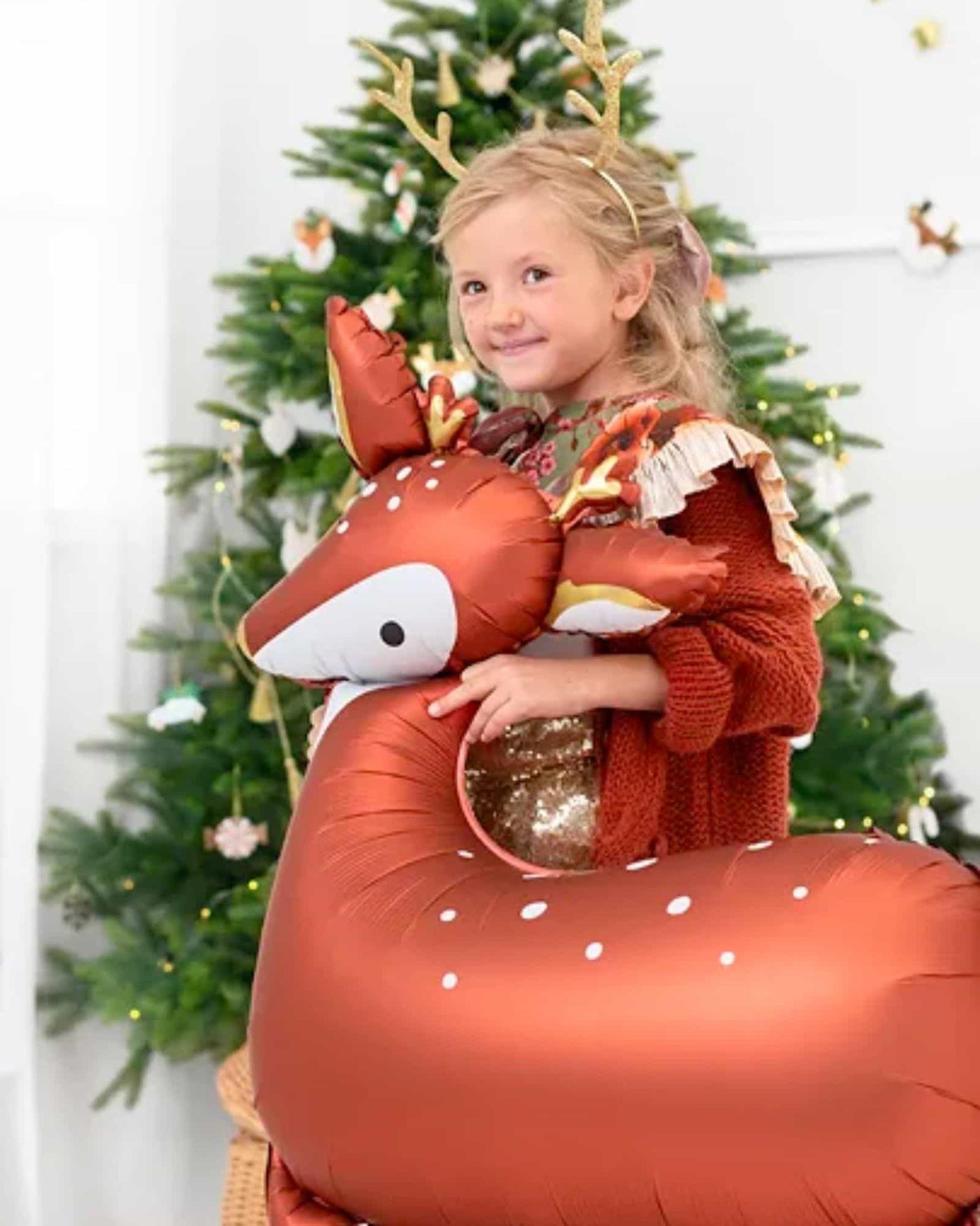 A child with golden antlers smiles while hugging a large, shiny, deer-shaped balloon near a decorated Christmas tree.