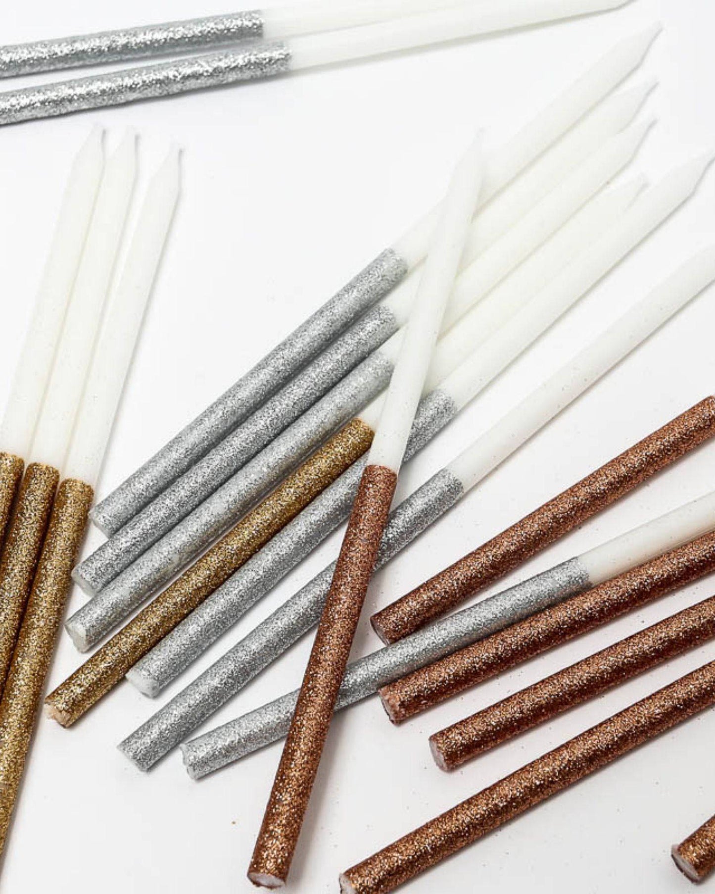 A collection of tall, glitter-dipped candles in silver, gold, and bronze hues against a white background.