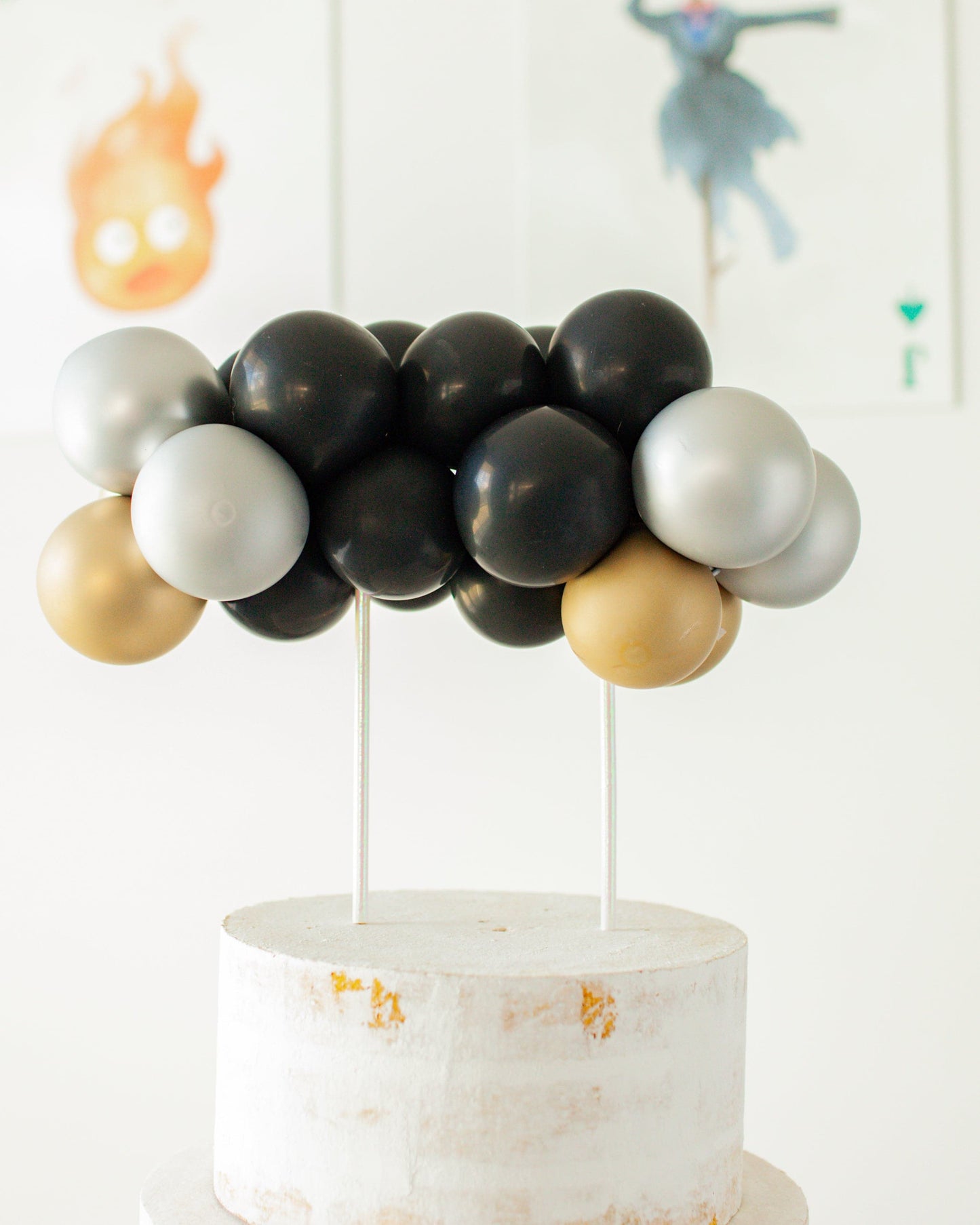 A cluster of black, silver, and gold balloons atop a rustic white cake stand, against a light background.