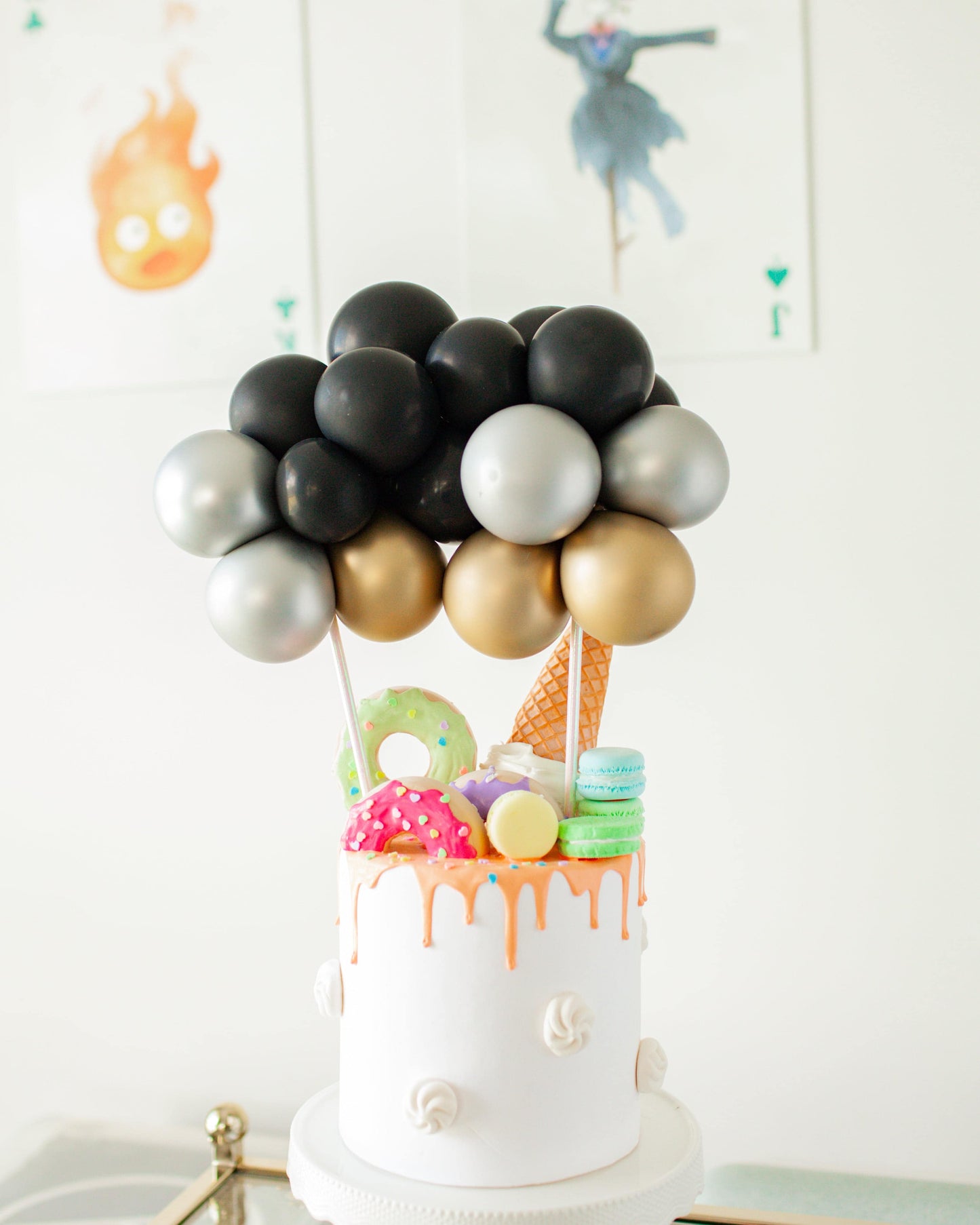 A colorful cake topped with shiny balloons and playful sweets, including donuts, macarons, and an ice cream cone.