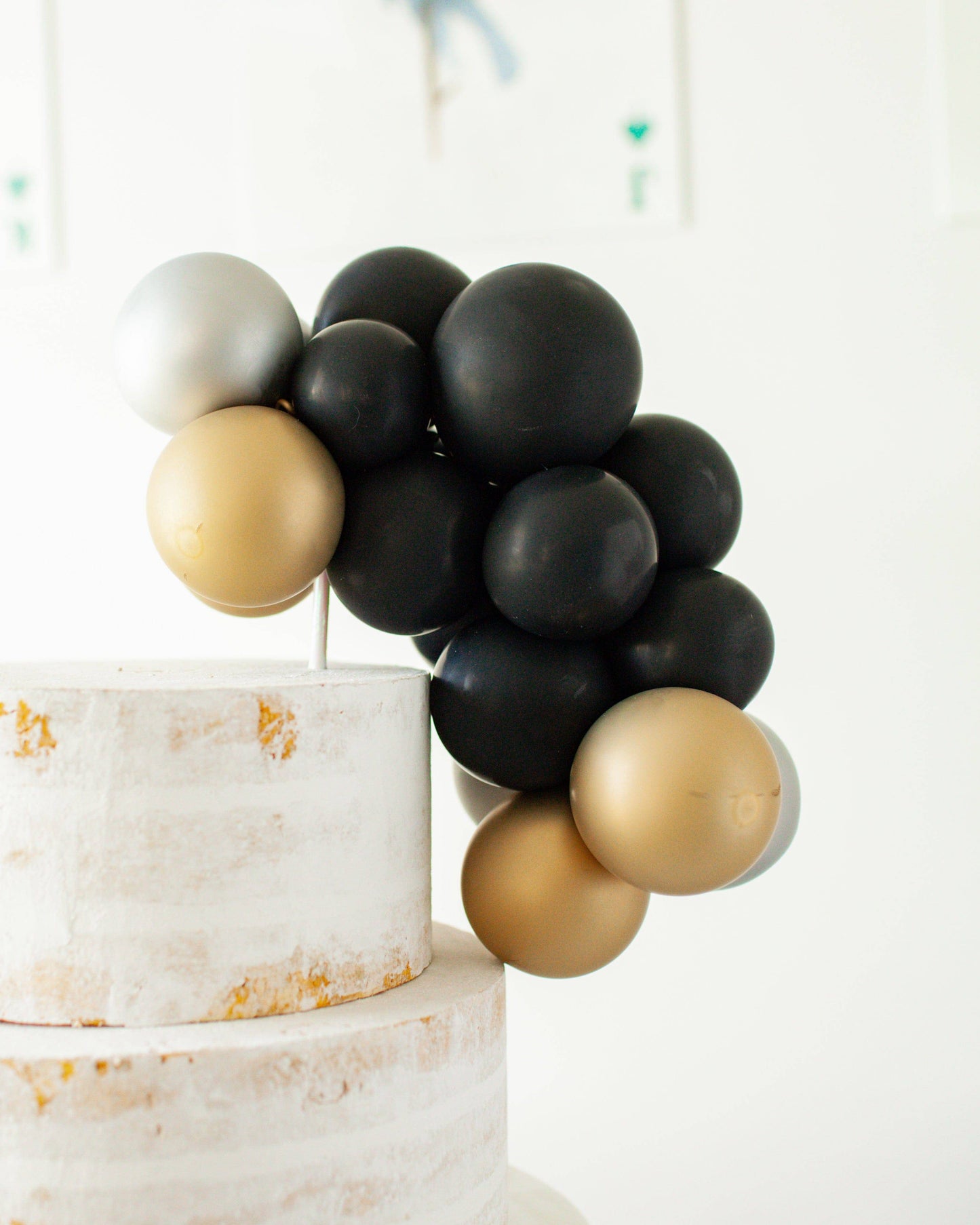 A cluster of black, gold, and silver balloons atop a textured white multi-tiered cake.
