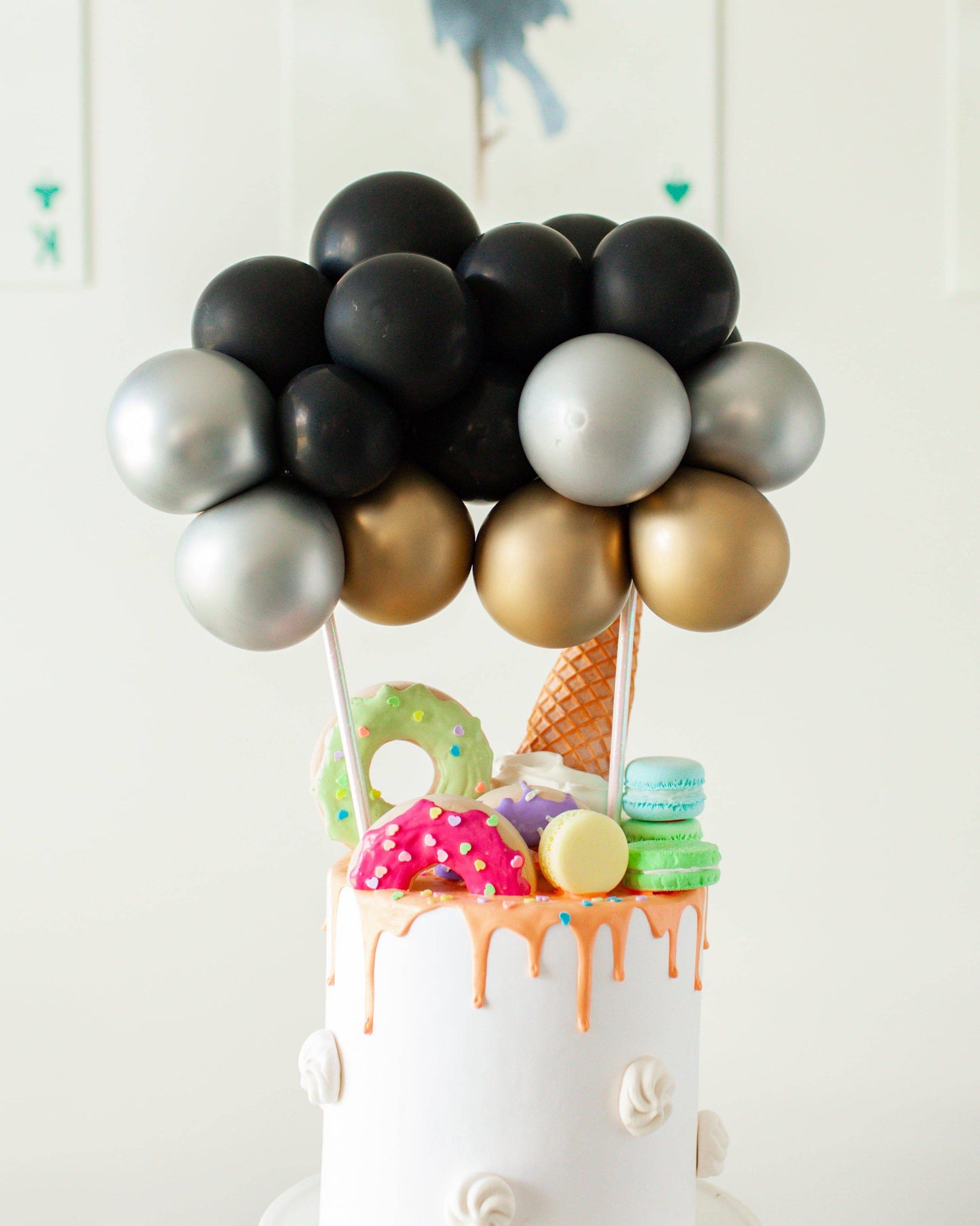 A whimsical cake topped with colorful balloons and playful sweets like donuts and macarons, perfect for celebrations.