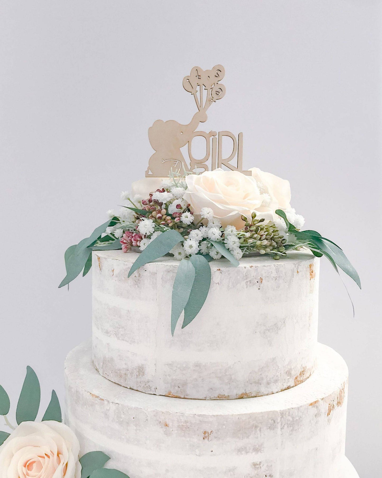 Wooden topper featuring a baby elephant and "girl," adorned with fresh flowers on a tiered, rustic cake.