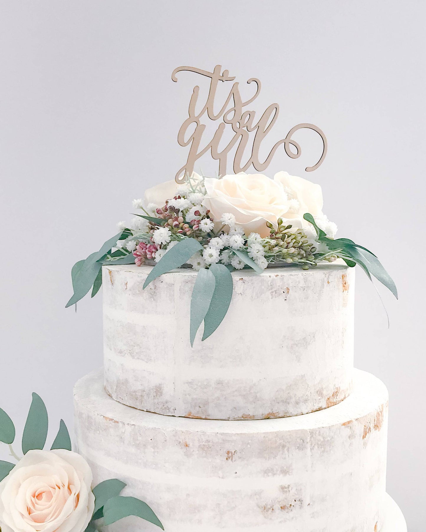 A white, layered cake adorned with a "It's a Girl" wooden topper and fresh roses and greenery.