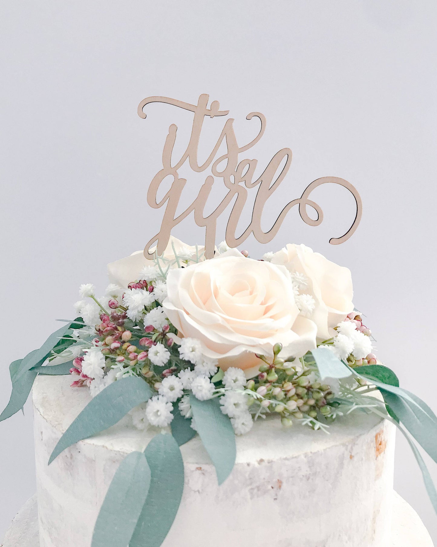 A wooden topper reads "It's a Girl" atop a cake adorned with soft pink roses and delicate greenery.