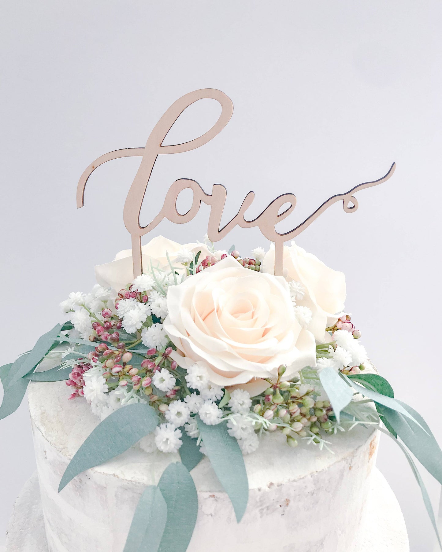 A wooden "love" topper on a floral arrangement of white roses and greenery, set against a soft background.