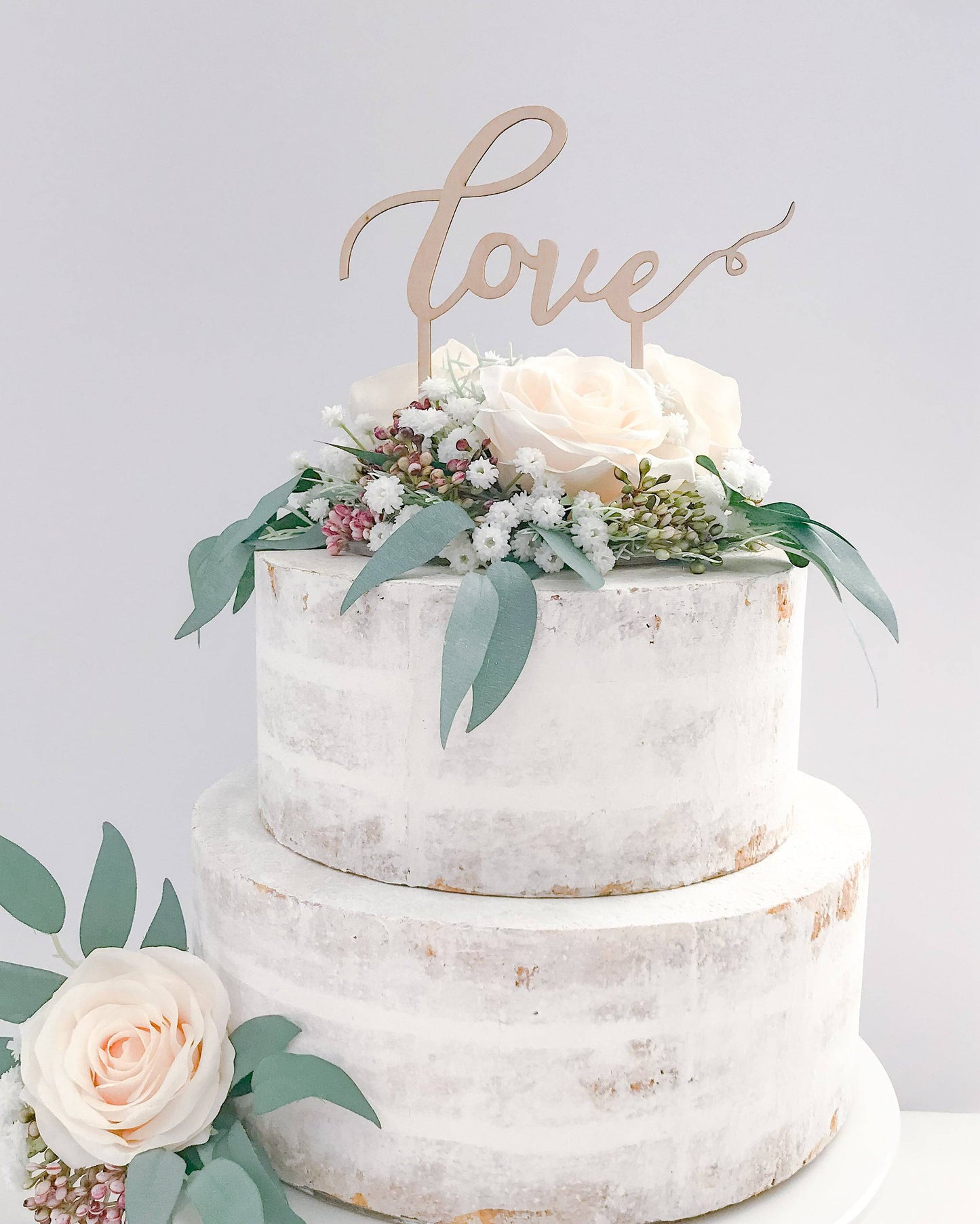 A tiered cake adorned with blush roses and greenery, topped with a wooden "love" sign against a light background.