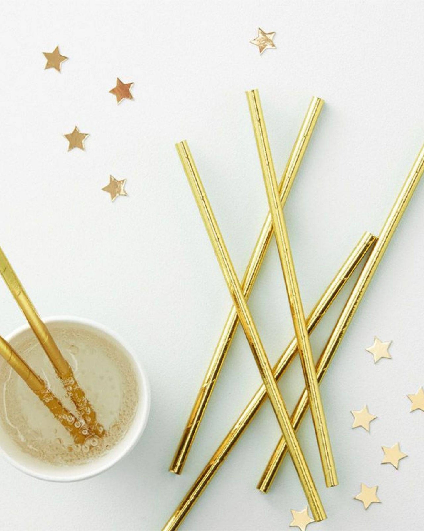 Gold foil straws arranged with a cup of bubbly drink and scattered star-shaped confetti on a light background.