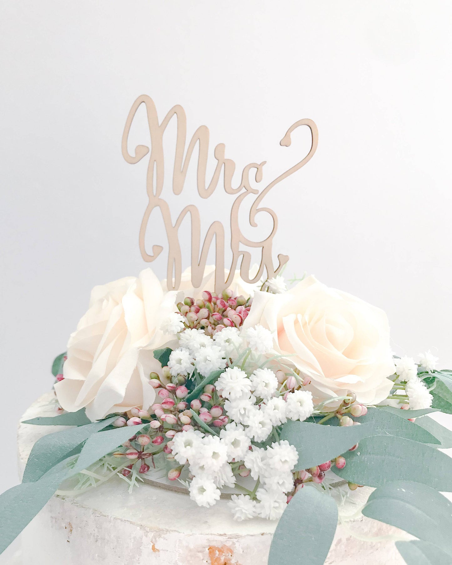 Elegant wooden topper with "Mr & Mrs" script atop soft pink roses and delicate white flowers, surrounded by greenery.