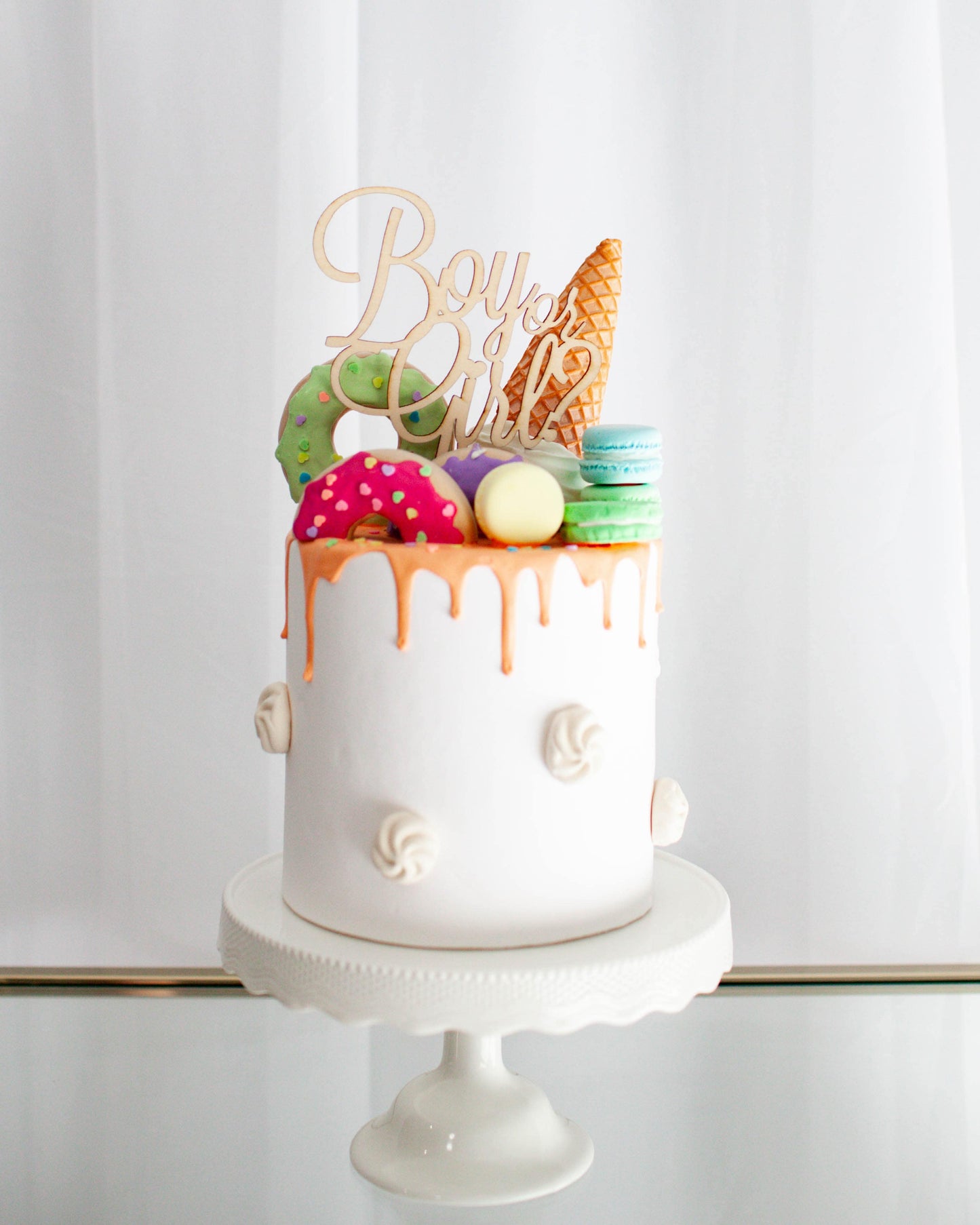 A colorful cake topped with a cone, donuts, and macarons, featuring a "Boy or Girl" sign in a bright setting.