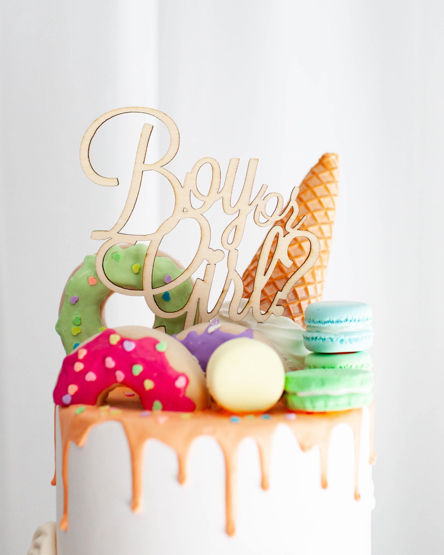 Colorful treats topped with a wooden sign reading "Boy or Girl?" adorn a white frosted cake with dripping orange icing.