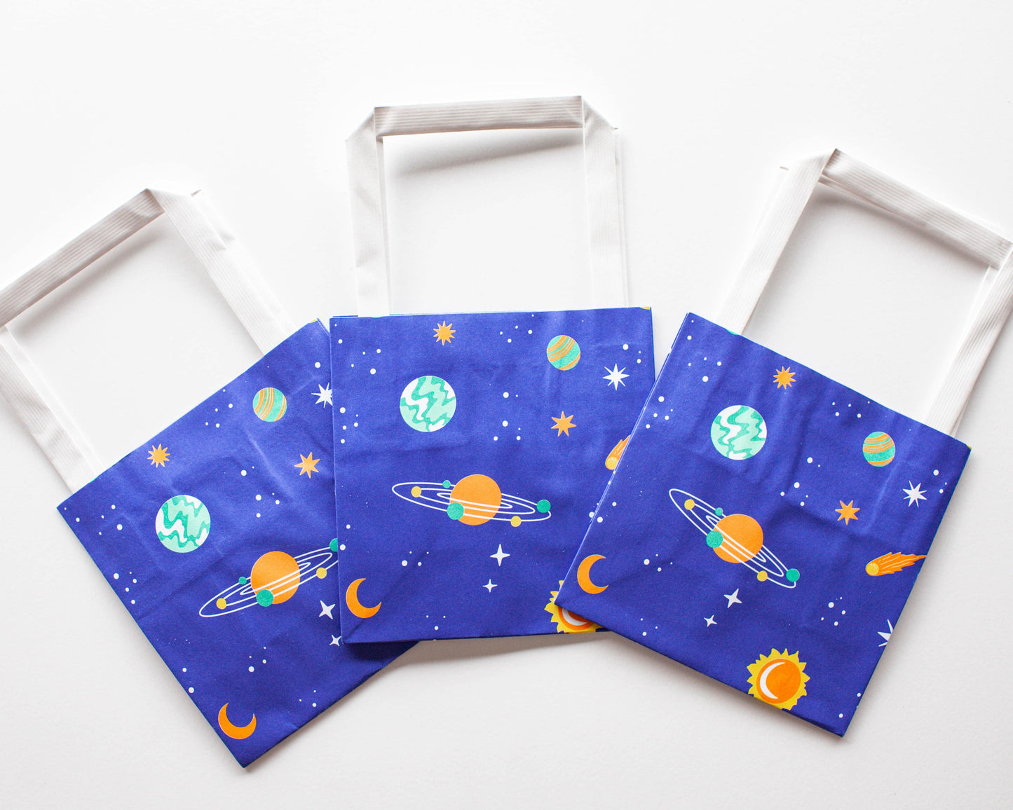 Three blue bags featuring colorful planets, stars, and celestial designs with white handles on a light background.