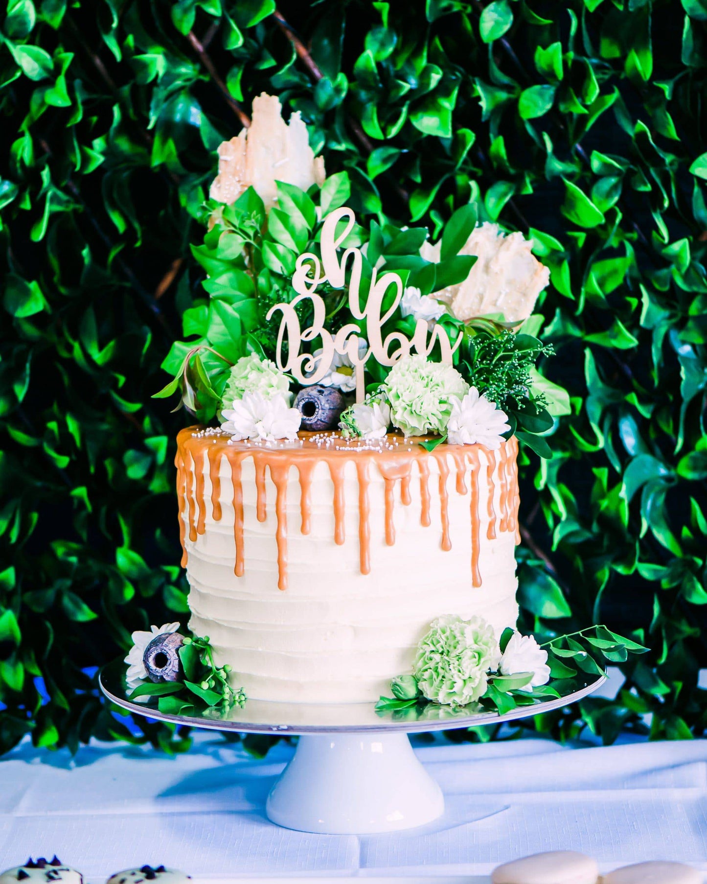 A tiered cake topped with greenery, flowers, and a wooden "oh baby" topper, set against a lush green backdrop.