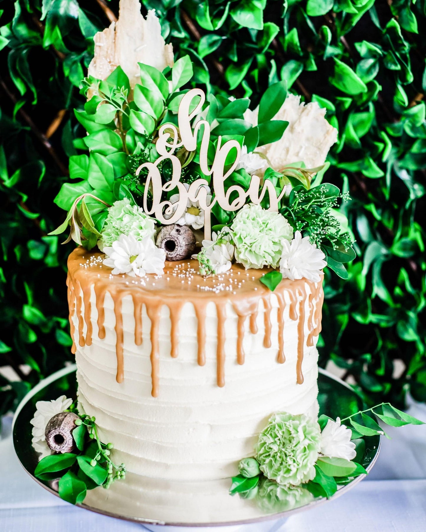 Layered cake topped with caramel drizzle, green and white flowers, and a wooden "Oh Baby" topper, set against leafy backdrop.