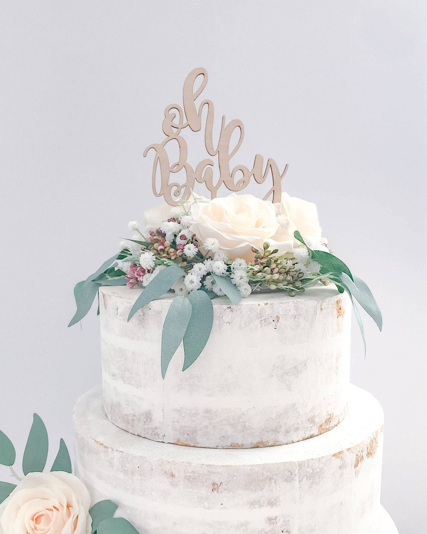 A rustic two-tier cake adorned with soft roses, greenery, and a wooden "oh baby" topper.