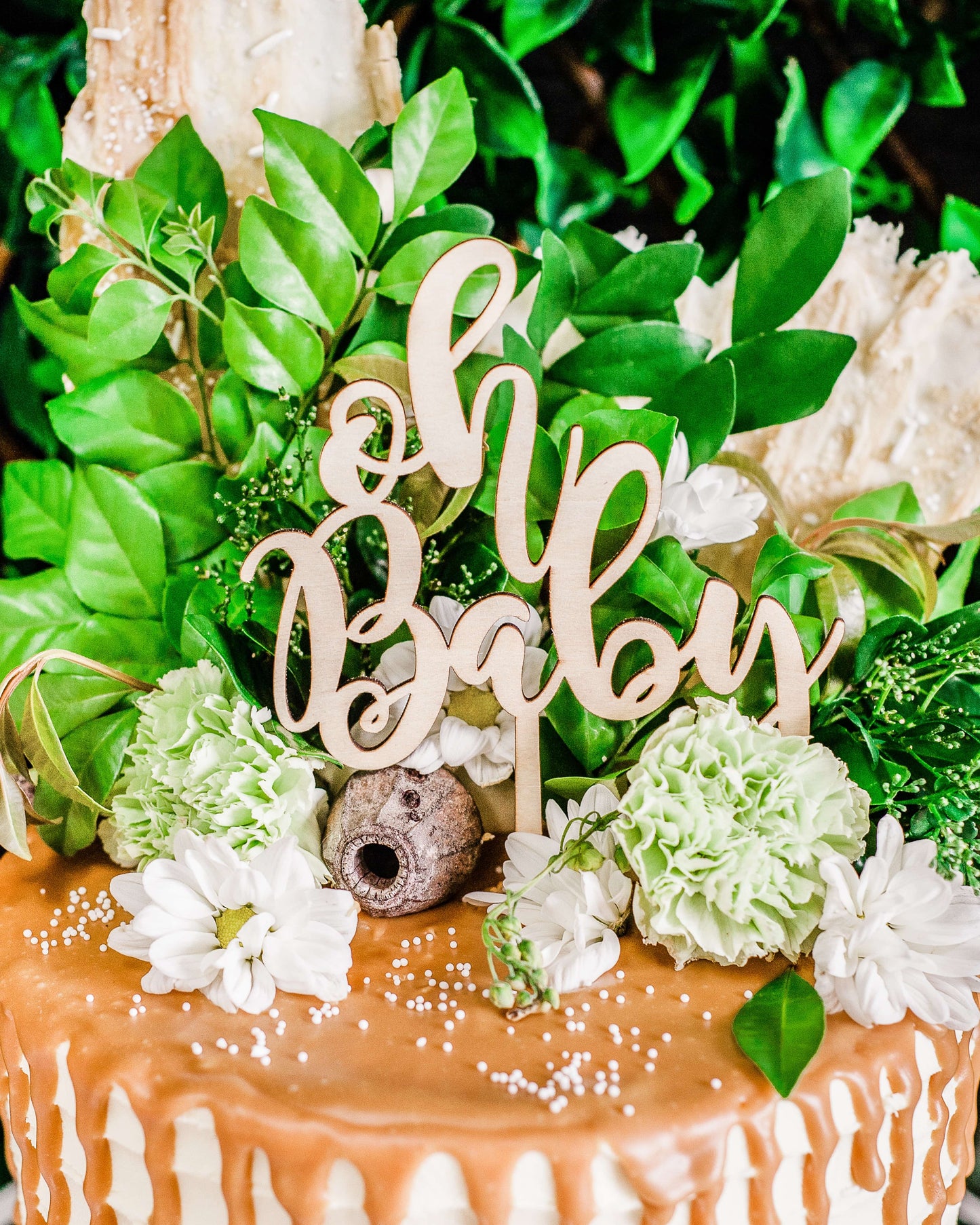 Wooden "Oh Baby" topper surrounded by greenery and white flowers on a decorated surface with caramel drizzle.