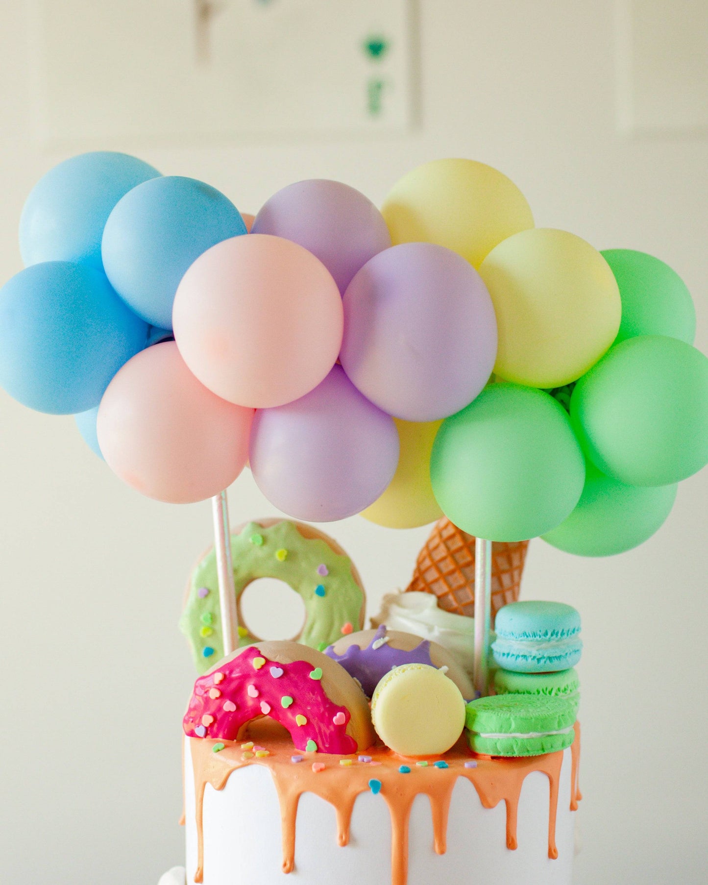Pastel balloons atop a colorful cake adorned with donuts, macarons, and dripping icing create a festive scene.