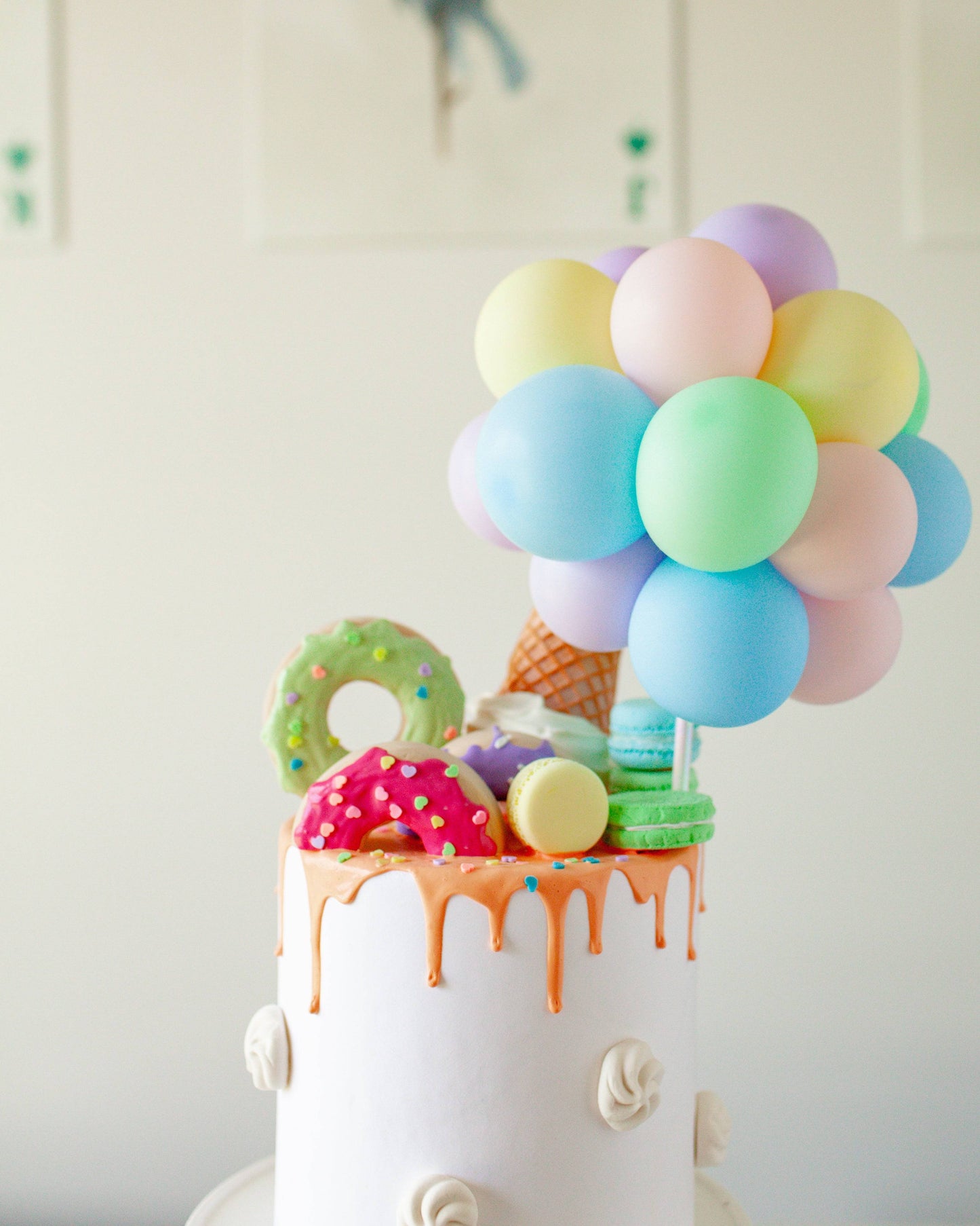 A pastel balloon cluster atop a white cake, adorned with colorful confections and dripping orange glaze.