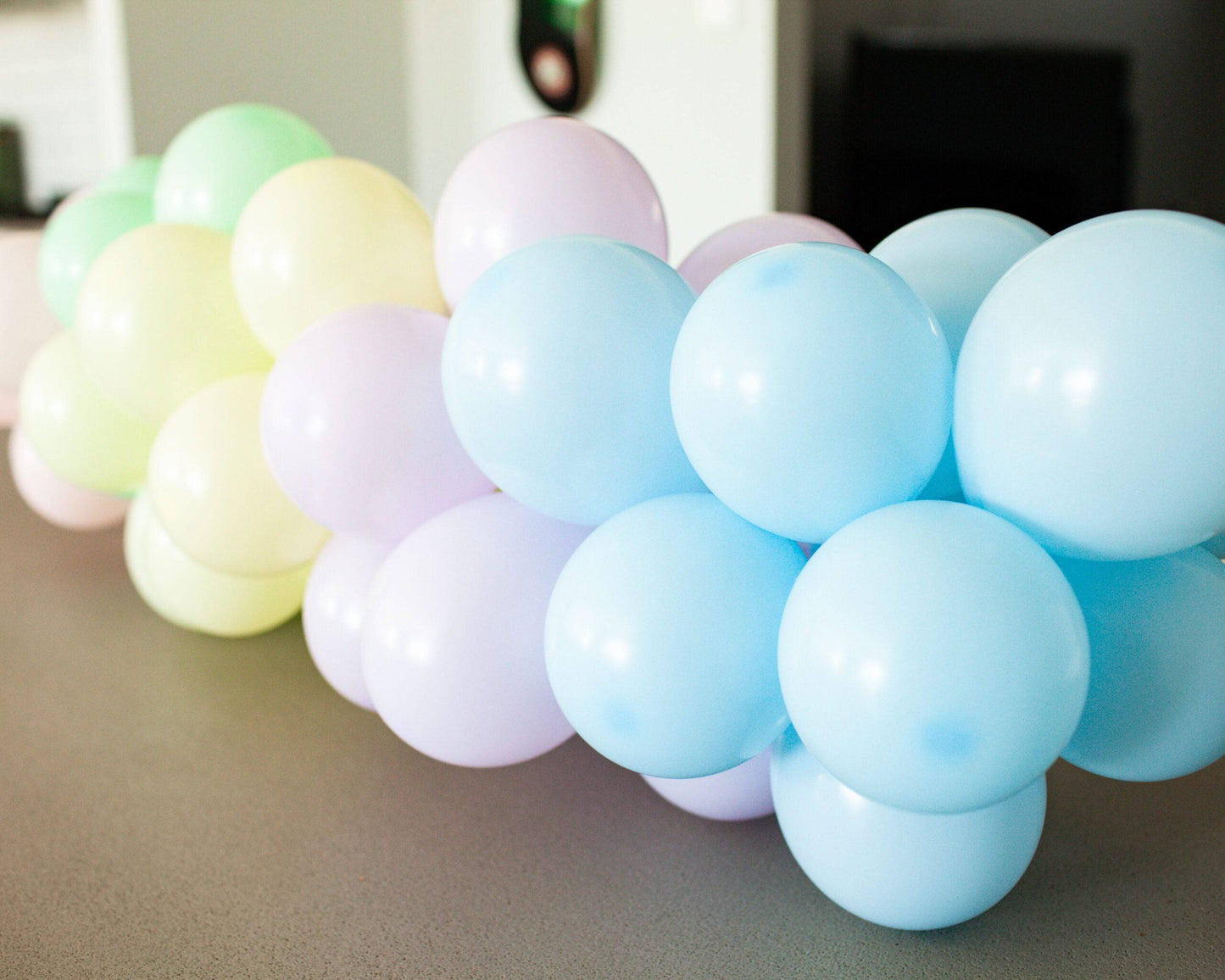 Pastel balloons in soft greens, yellows, blues, and purples arranged in a playful cluster on a grey surface.