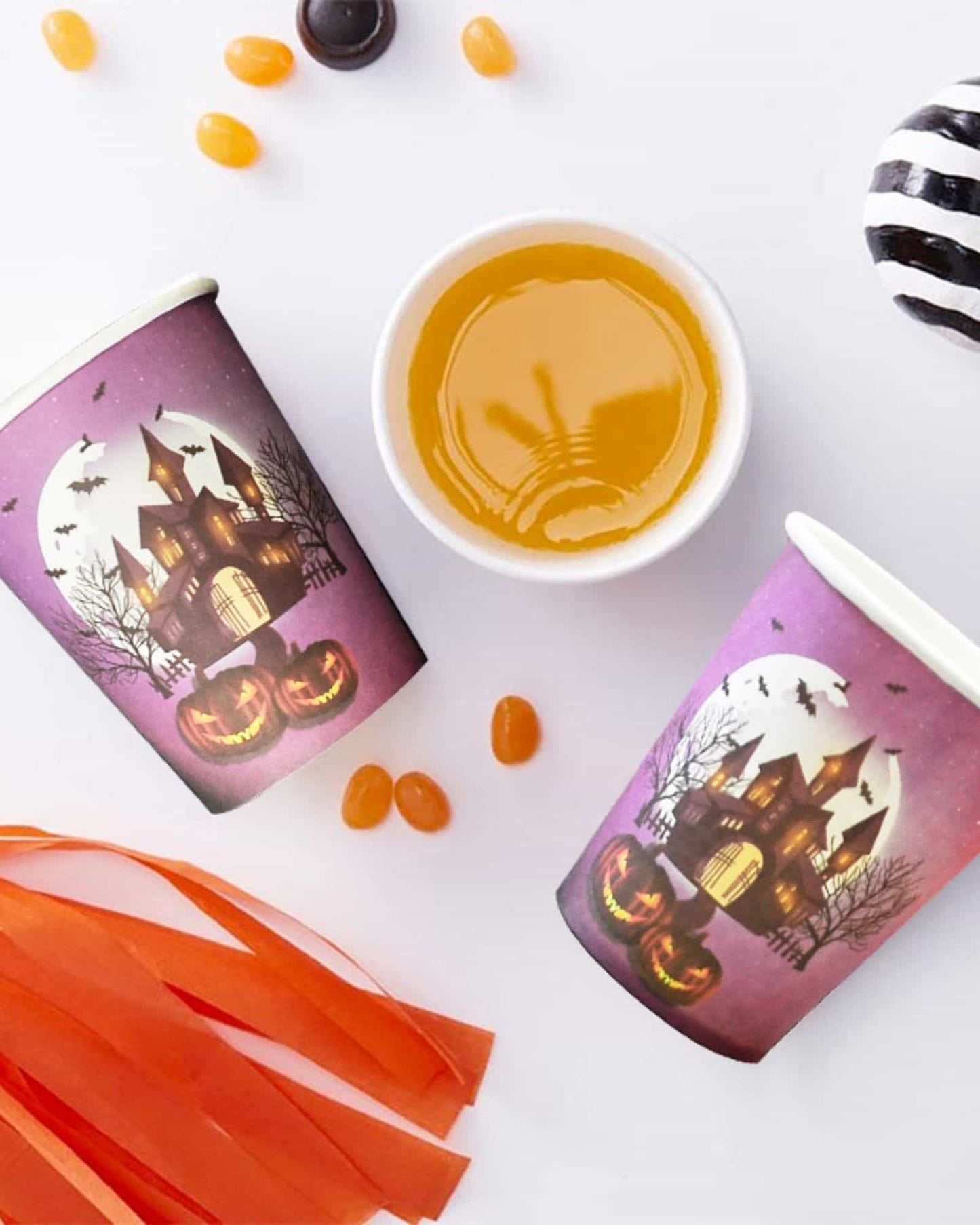 Two Halloween-themed cups feature a spooky castle and glowing jack-o'-lanterns, surrounded by candy and orange strips.