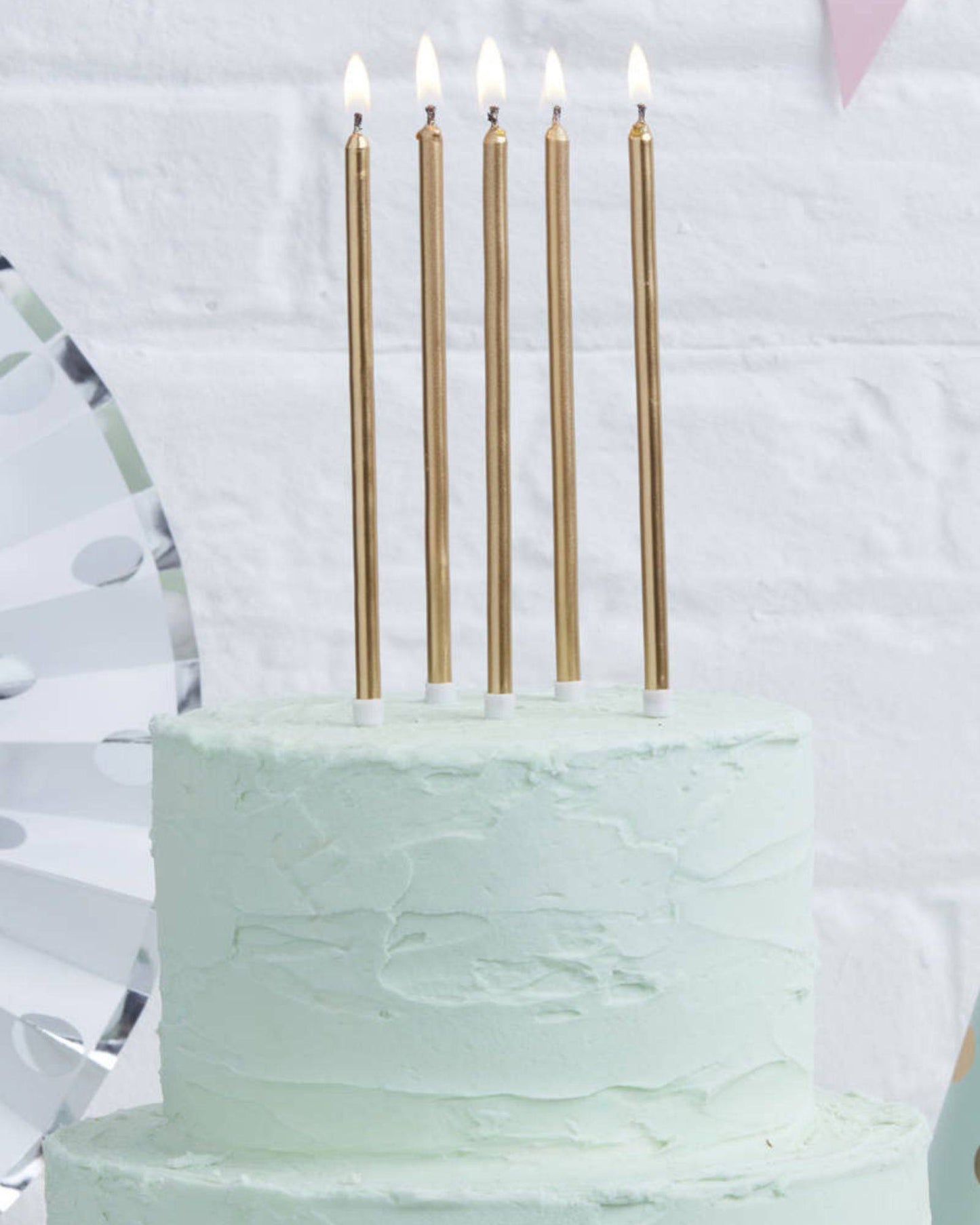Five tall, gold candles with flames atop a pastel green cake, set against a light-colored backdrop.