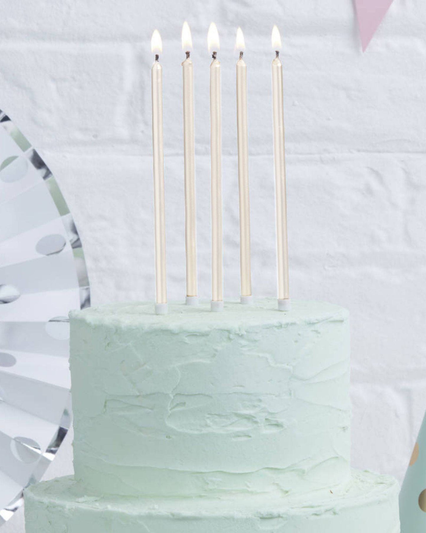 Five tall, pearl white candles with lit flames atop a light green frosted cake against a textured white background.
