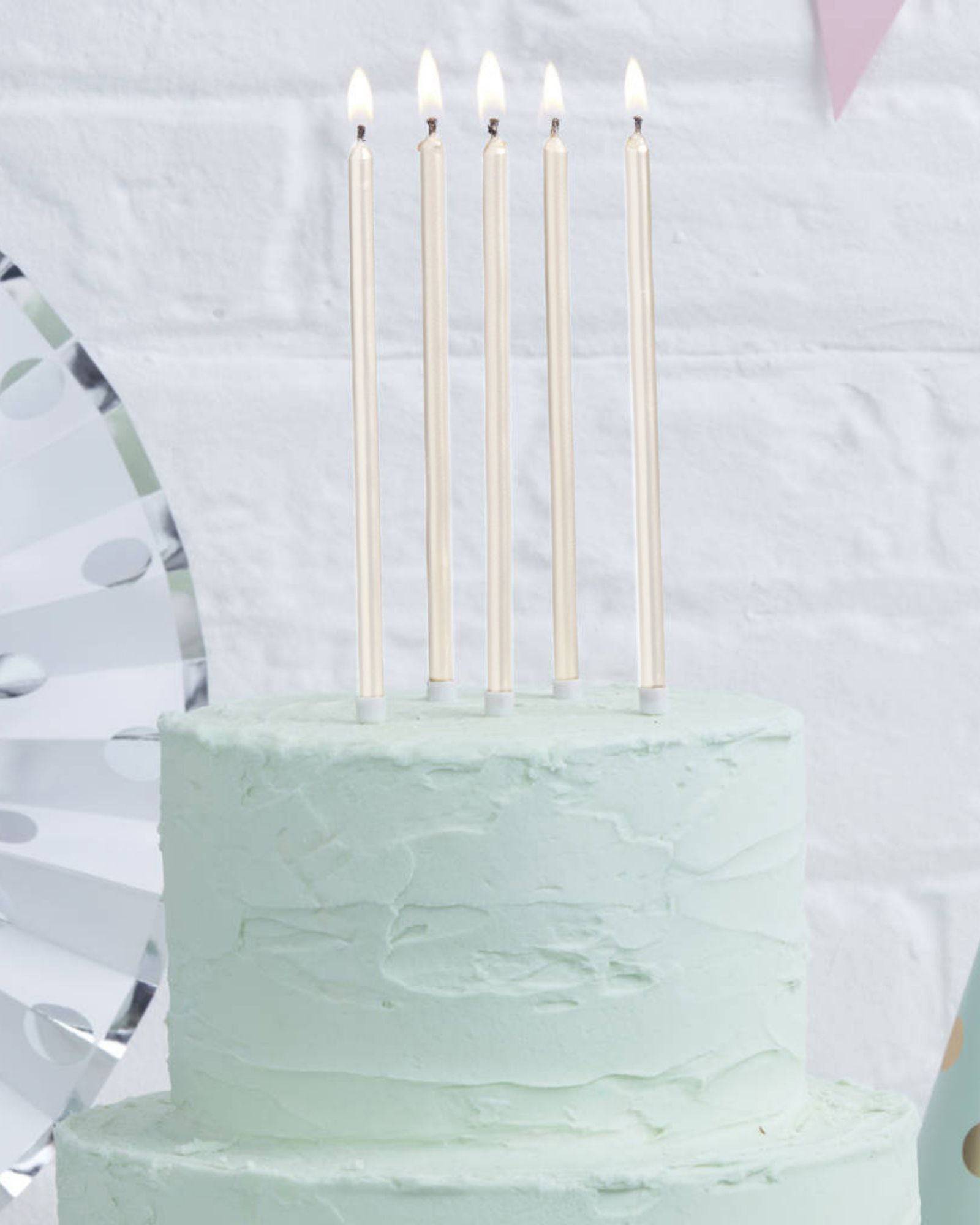 Five tall, pearl white candles with lit flames atop a light green frosted cake against a textured white background.