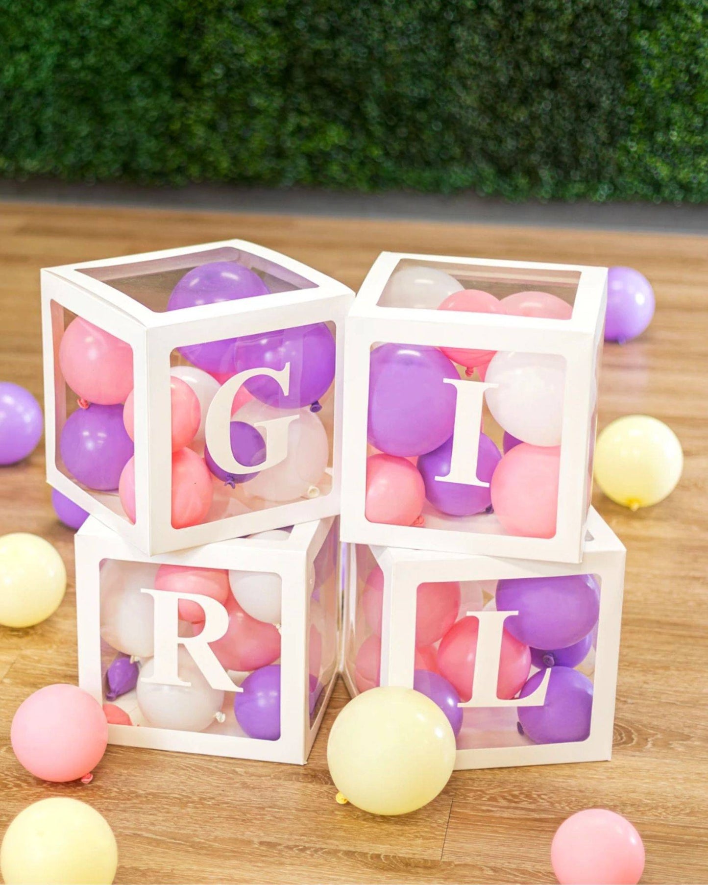 Four clear boxes filled with colorful balloons, displaying the letters G, I, R, and L, on a wooden floor.