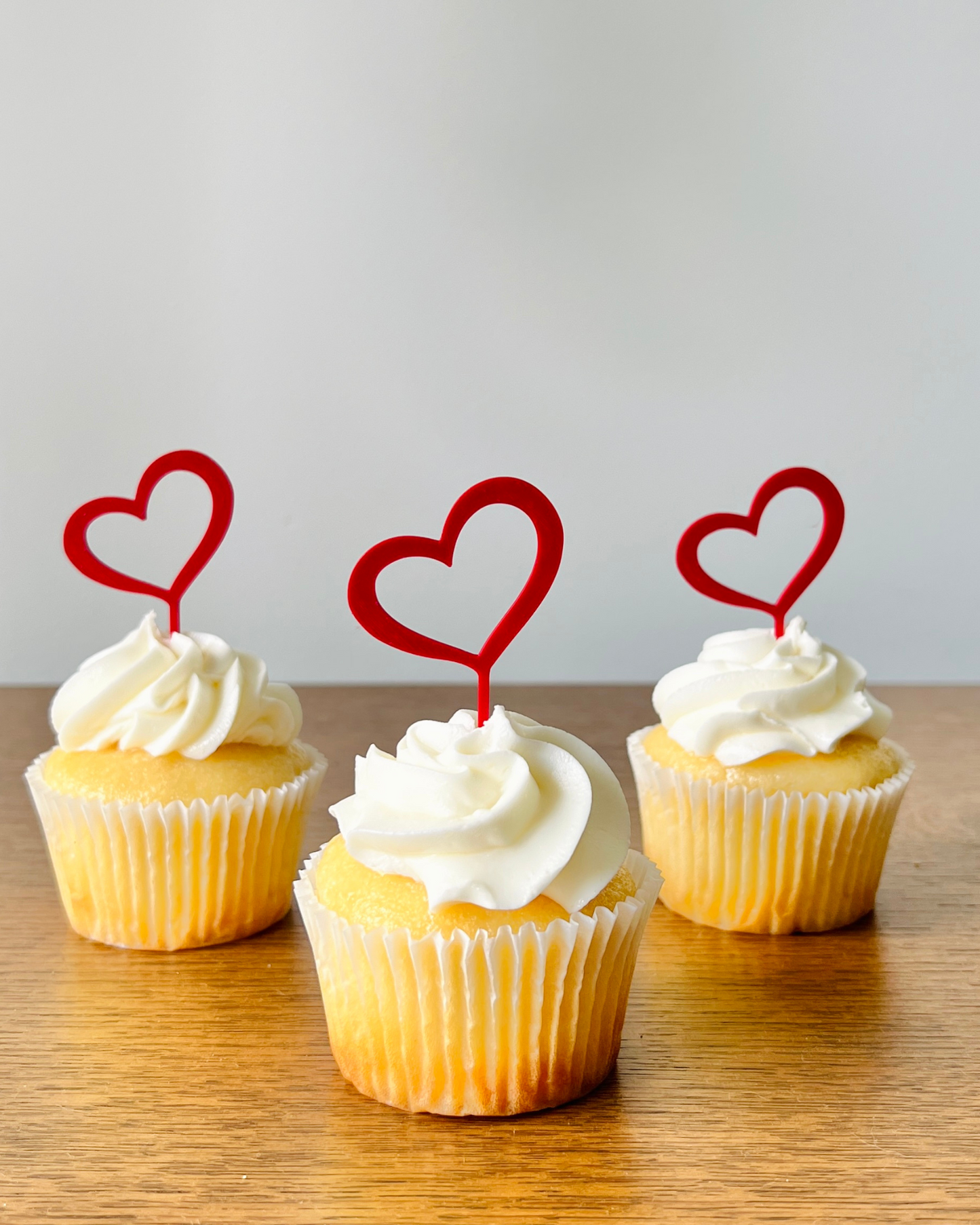 Three yellow cupcakes topped with swirls of white frosting and red heart-shaped picks, set against a light background.