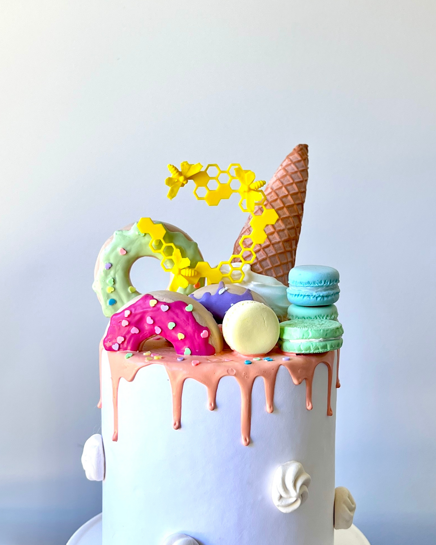 A colorful cake topped with playful decorations like donuts, macarons, and an ice cream cone, drizzled with icing.