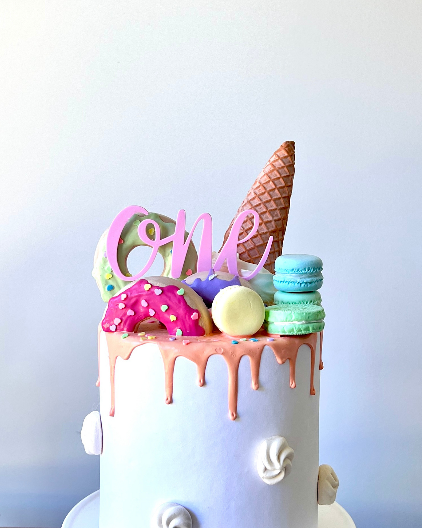 Colorful cake topped with a cone, macarons, and a donut, featuring a pink "ONE" sign with dripping icing.