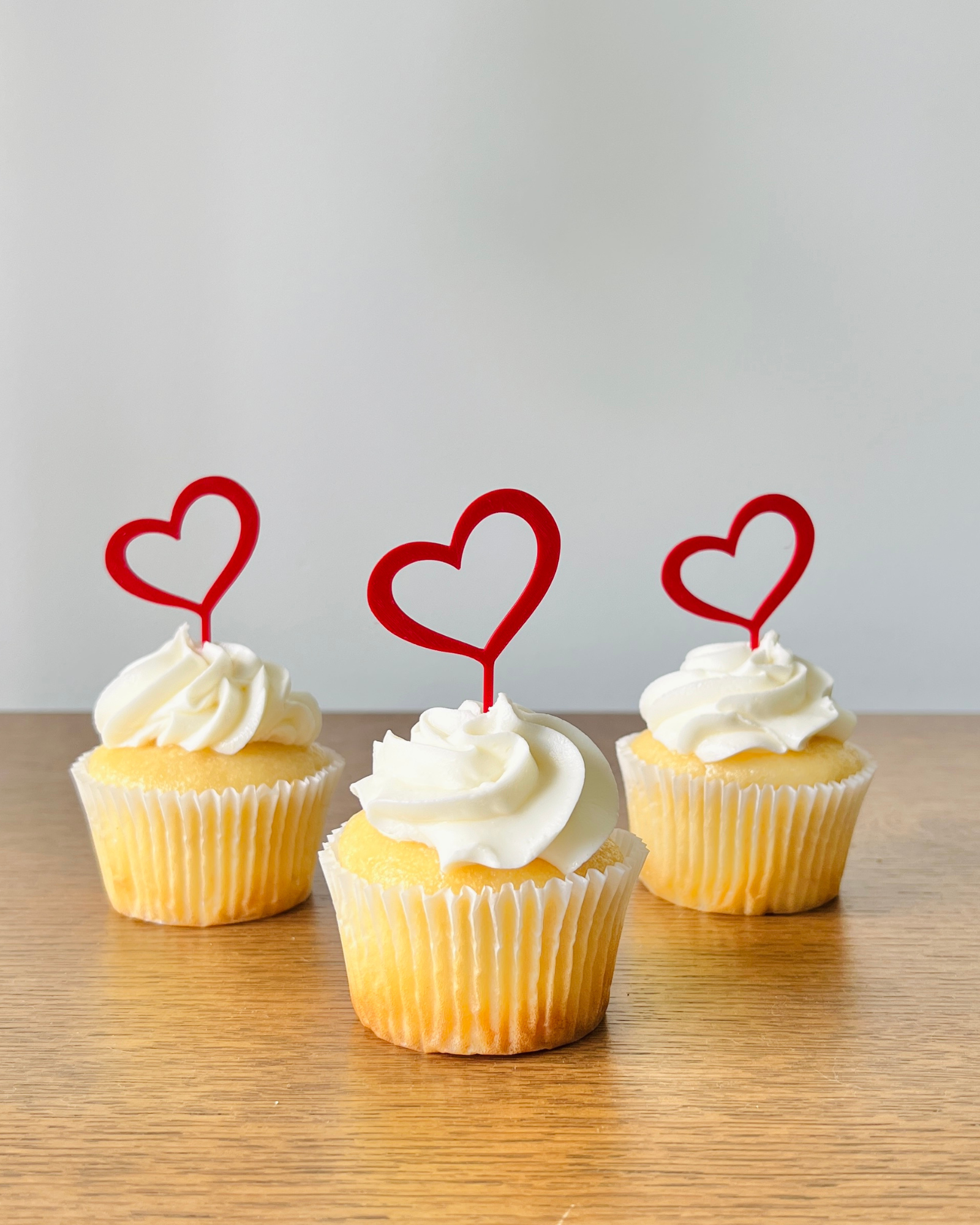 Three yellow cupcakes topped with white frosting and red heart-shaped decorations on wooden surface.