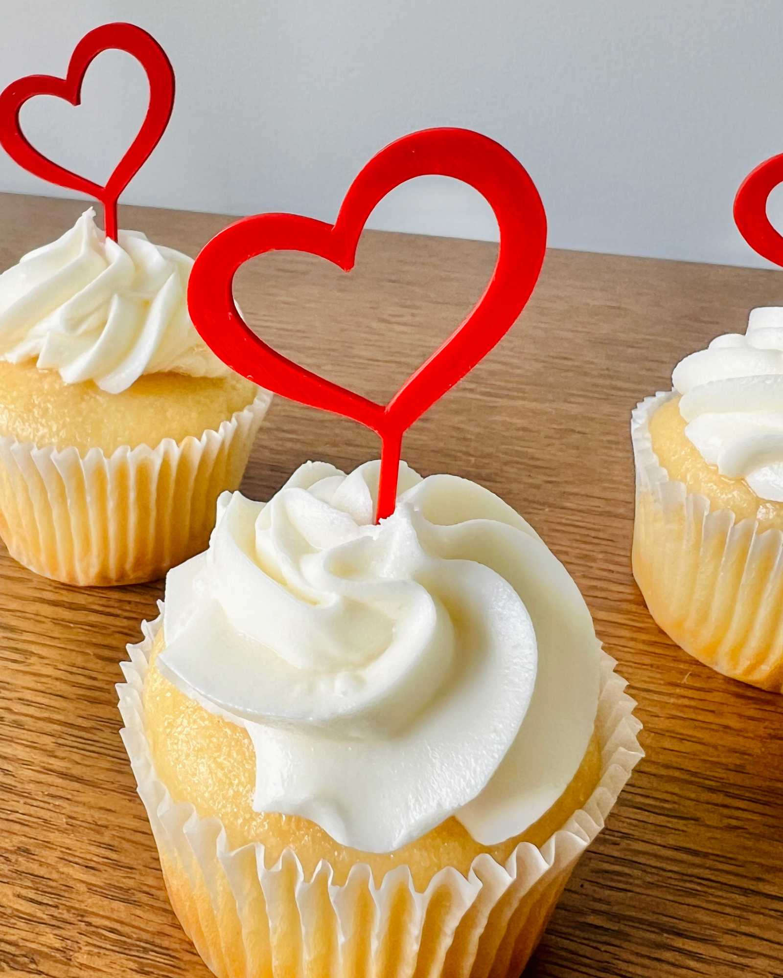 Three vanilla cupcakes topped with swirls of white frosting and red heart-shaped decorations, set on a wooden surface.