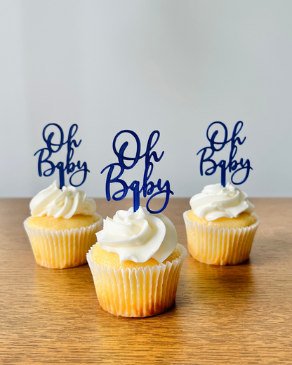 Three yellow cupcakes topped with swirls of white frosting and decorated with blue "Oh Baby" toppers on a wooden surface.