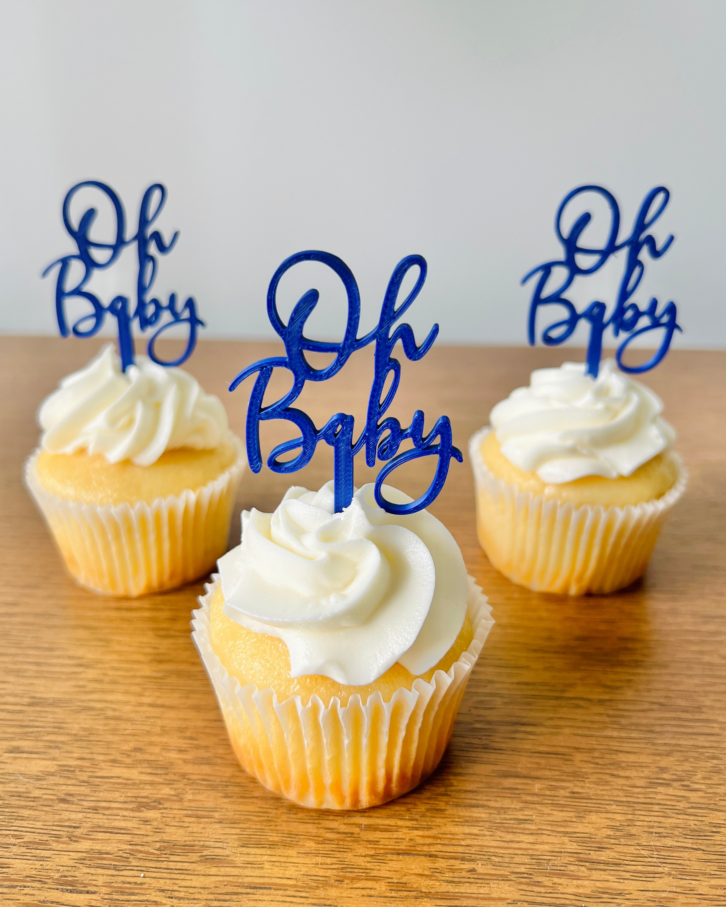 Three yellow cupcakes with white frosting topped with blue "Oh Baby" cake toppers on a wooden surface.