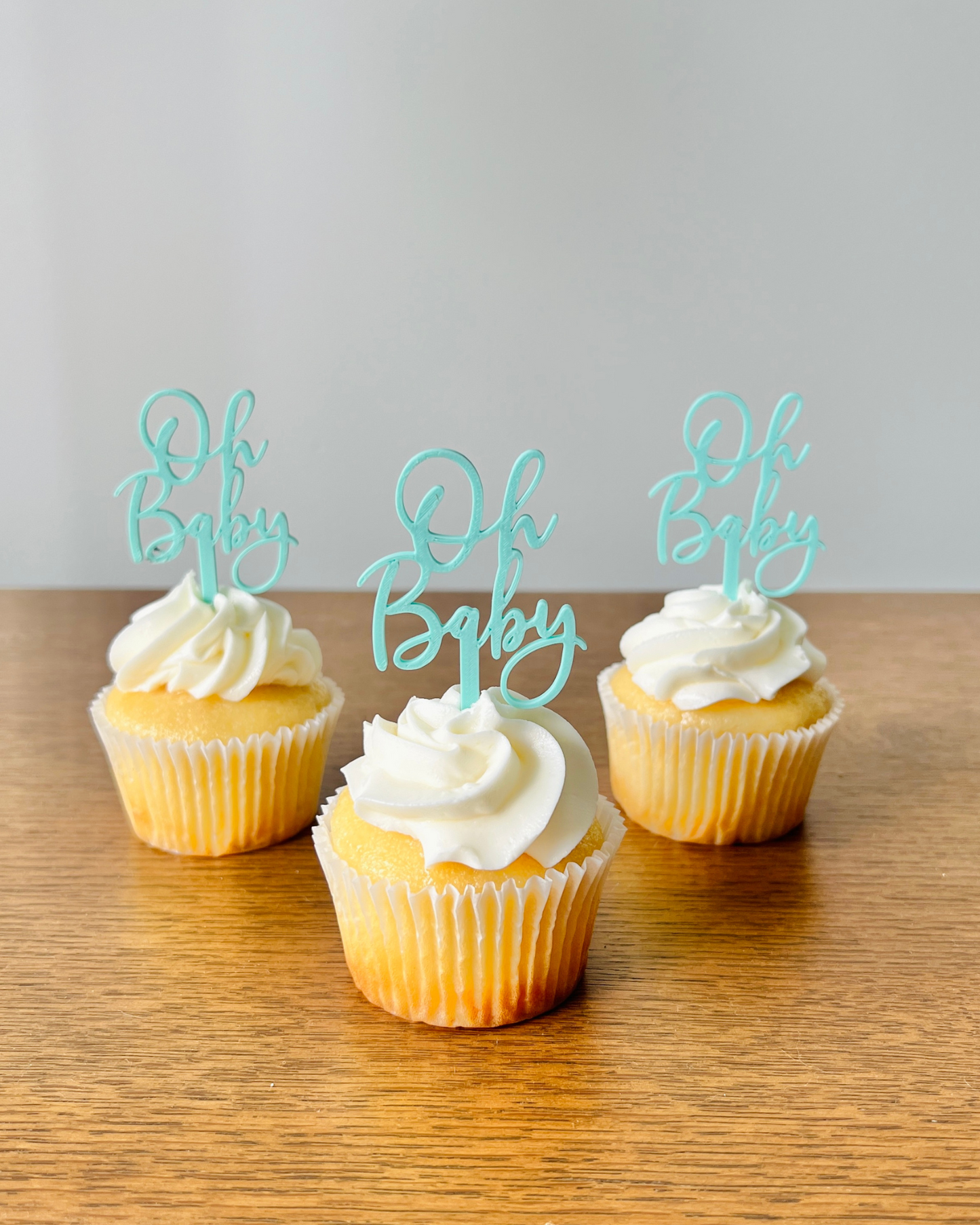 Three yellow cupcakes topped with white frosting and aqua "Oh Baby" toppers on a wooden surface.