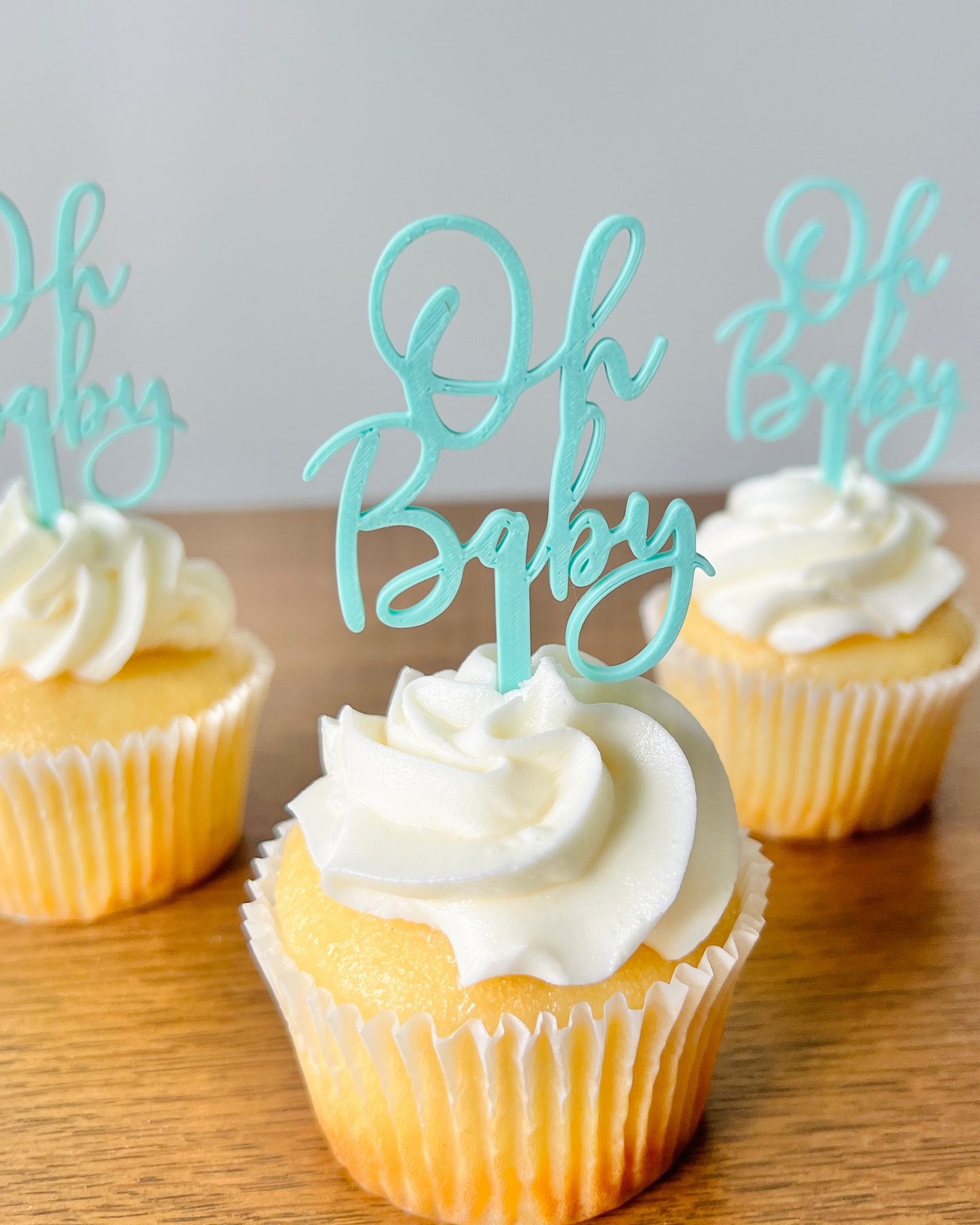 Three yellow cupcakes topped with white frosting and blue "Oh Baby" toppers on a wooden surface.