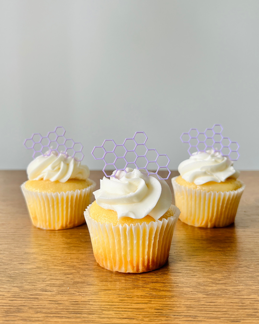 Three yellow cupcakes with swirls of white frosting topped with purple honeycomb decorations on a wooden surface.