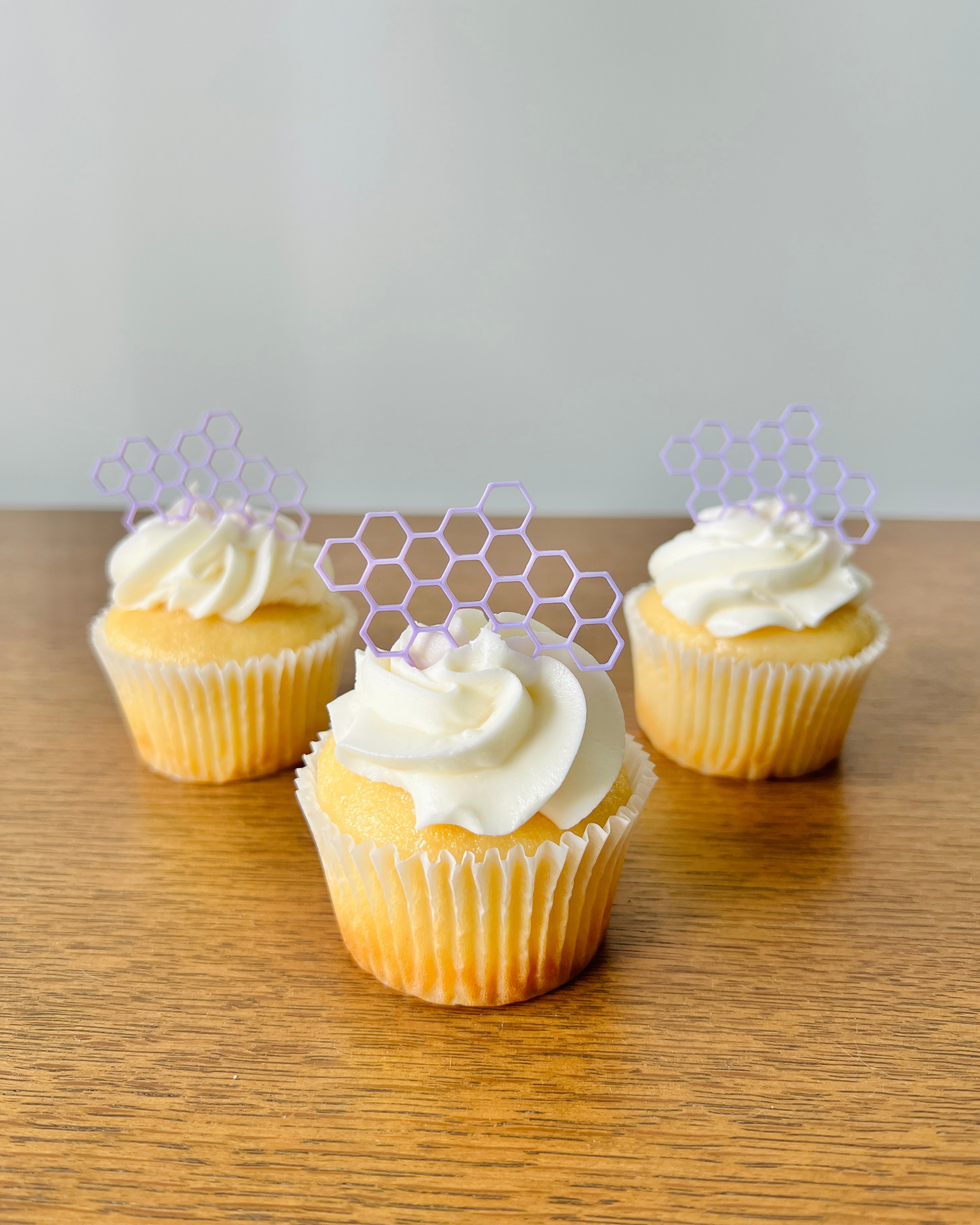 Three yellow cupcakes with swirls of white frosting, topped with purple honeycomb decorations, on a wooden surface.