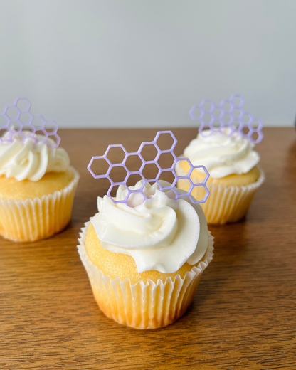 Three yellow cupcakes with swirls of white frosting topped with purple honeycomb-shaped decorations on a wooden surface.