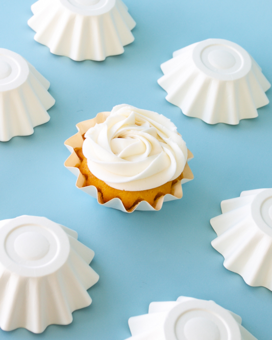 A golden treat topped with swirls of white frosting, surrounded by white fluted molds on a blue backdrop.