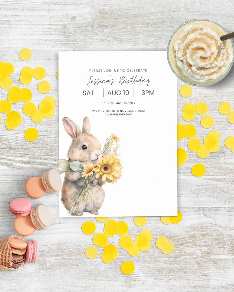 A cute rabbit holds a bouquet of sunflowers beside colorful macarons and yellow confetti on a wooden surface.